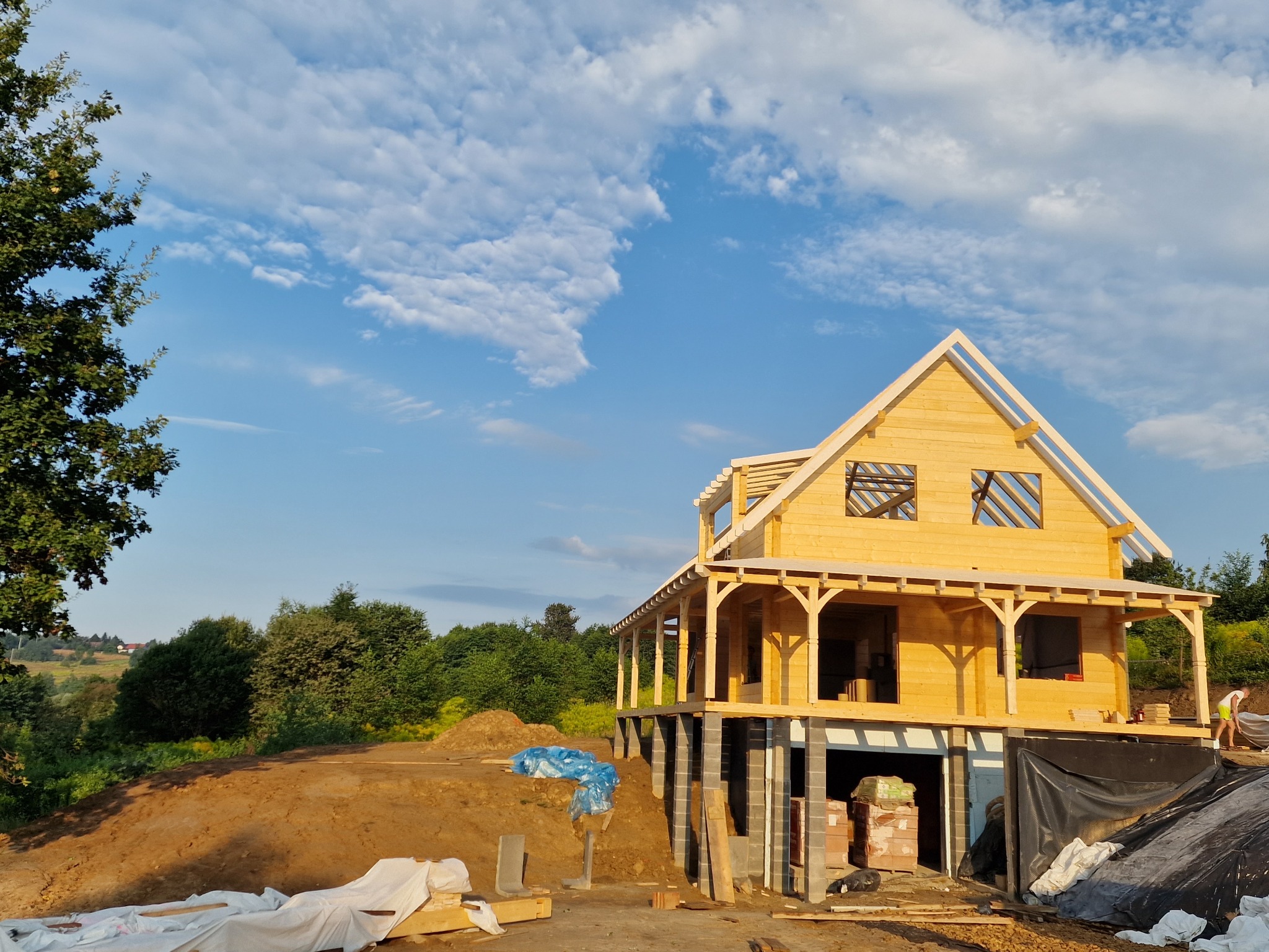 Wznoszenie domu szkieletowego na fundamencie z bloczków betonowych, widok z dołu na konstrukcję w surowym stanie, błękitne niebo w tle.