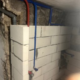 Hydro Atak - Instalacja wodna z rurami w kolorze niebieskim i czerwonym, zamontowana na ścianie z białych bloczków. Widoczne fragmenty starej, kamiennej ściany.