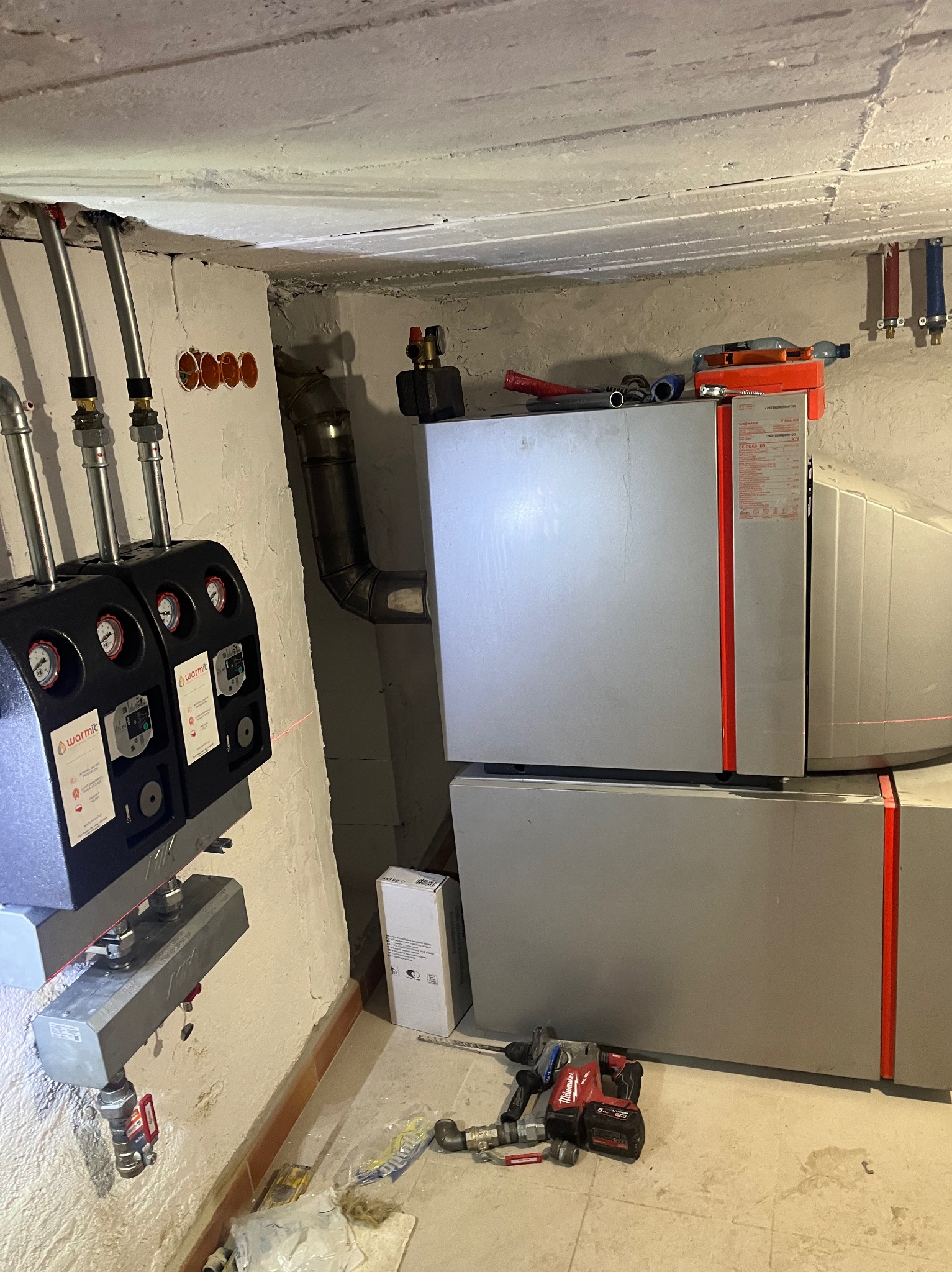 Szary piec CO z czarnymi elementami sterującymi, rurami i narzędziami na podłodze w piwnicy. Widoczne przewody i instalacje.