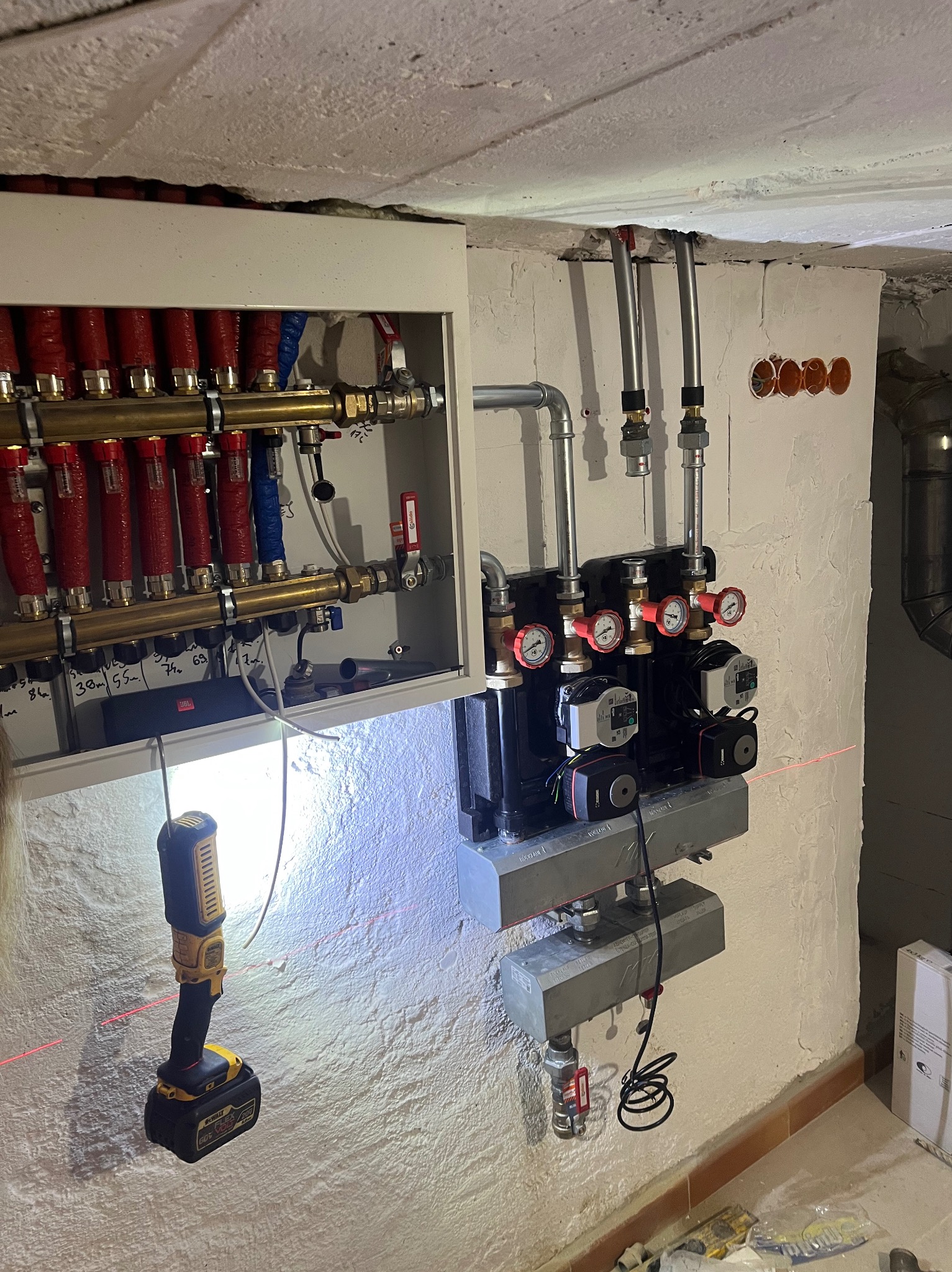 Rozdzielacz ogrzewania podłogowego z pompami obiegowymi, manometrami i rurami. Widoczny laserowy niwelator i narzędzia. Biała ściana z surowym wykończeniem.