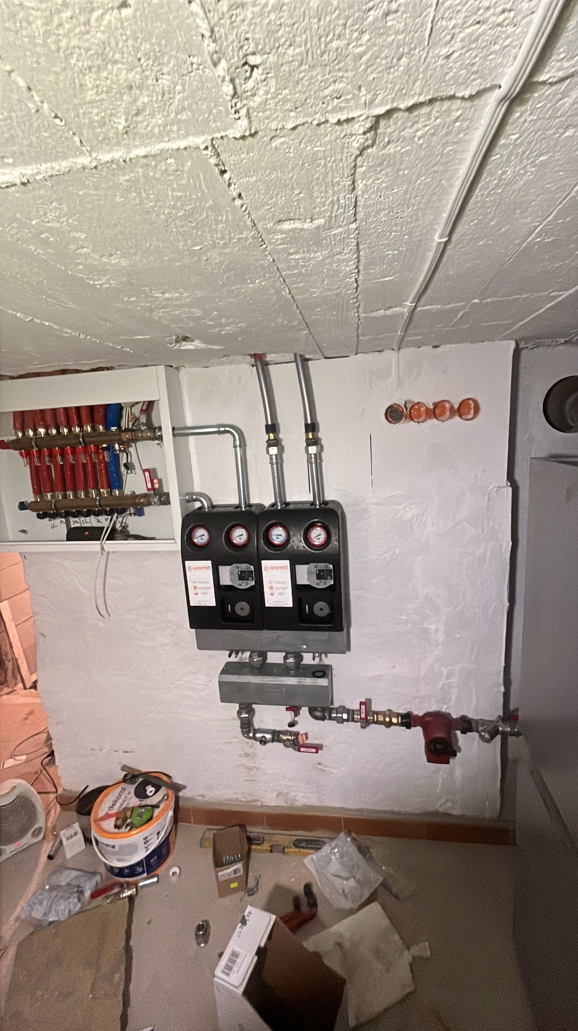 Instalacja rozdzielacza ogrzewania podłogowego Warmit z manometrami, rurami i pompą obiegową na tle surowej ściany. Na podłodze widoczne narzędzia i materiały instalacyjne.