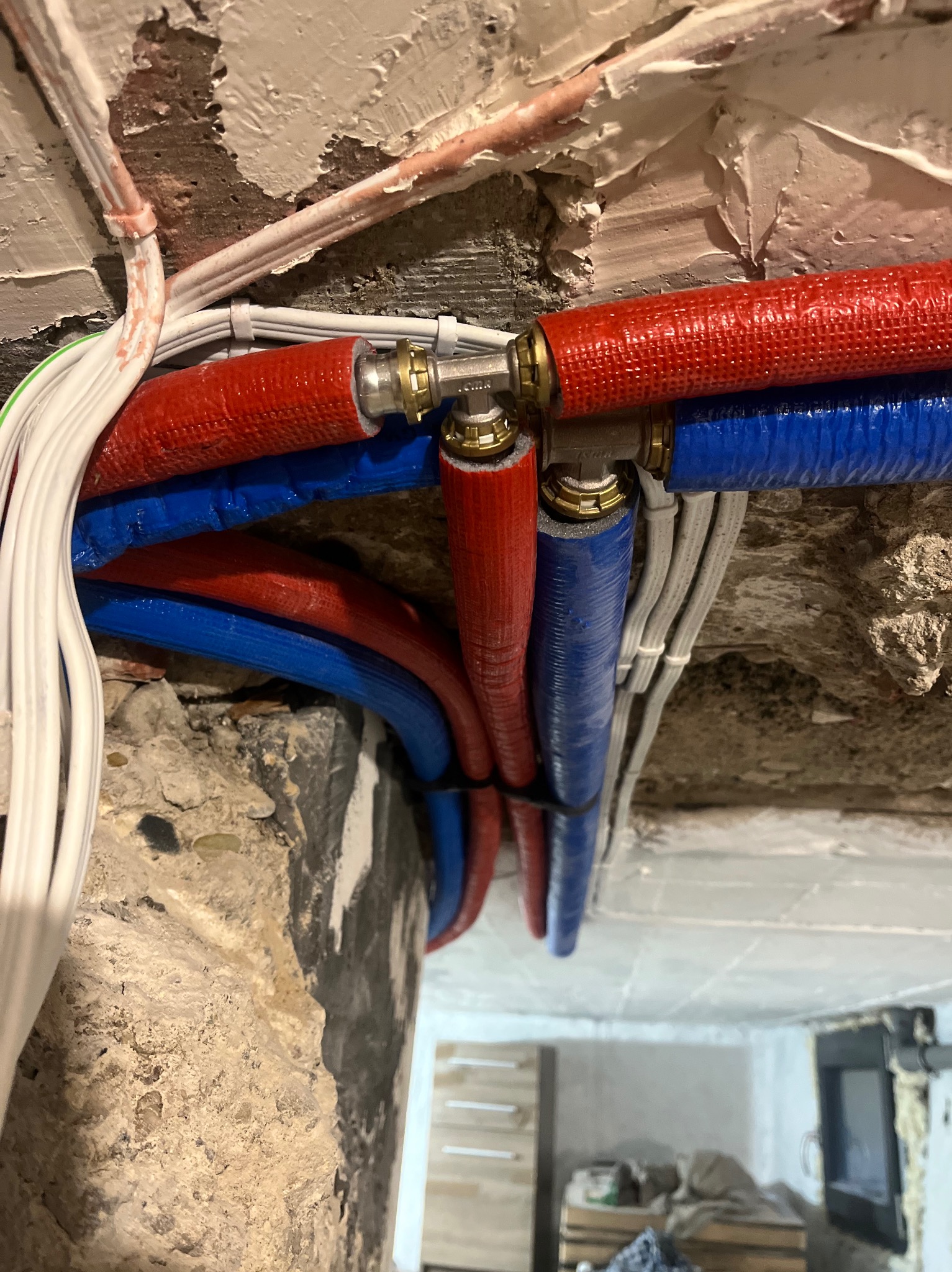 Instalacja wodna z rur PEX w kolorach czerwonym i niebieskim, z mosiężnymi złączkami, zamocowana do surowego muru. Widoczne także przewody elektryczne.