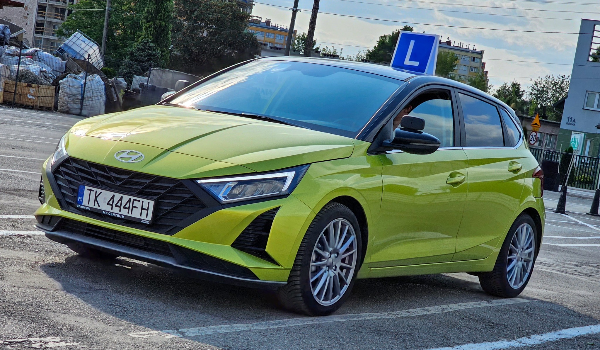 Limonkowy Hyundai i20 z niebieską literą 'L' na dachu, zaparkowany na placu manewrowym. Widoczne budynki w tle i oznaczenia poziome na asfalcie.