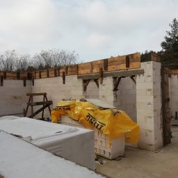 AURABET SPÓŁKA Z OGRANICZONĄ ODPOWIEDZIALNOŚCIĄ - Wznoszenie ścian z bloczków betonu komórkowego Ytong. Widoczne drewniane szalunki nad otworami okiennymi i drzwiowymi. Część bloczków przykryta żółtą folią.