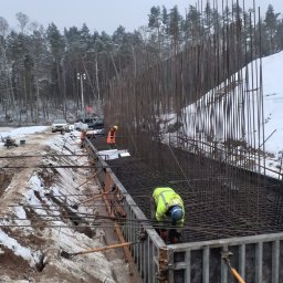 AURABET SPÓŁKA Z OGRANICZONĄ ODPOWIEDZIALNOŚCIĄ - Zbrojenie fundamentów pod budowę w zimowej scenerii. Widoczni pracownicy w odblaskowych ubraniach i stalowe pręty zbrojeniowe tworzące konstrukcję.