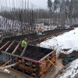 AURABET SPÓŁKA Z OGRANICZONĄ ODPOWIEDZIALNOŚCIĄ - Zbrojenie i szalunek pod fundamenty mostu w zimowej scenerii. Widoczne pręty zbrojeniowe, drewniane podpory i pracownicy w odblaskowych ubraniach.