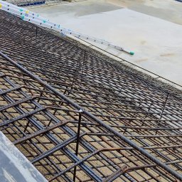 AURABET SPÓŁKA Z OGRANICZONĄ ODPOWIEDZIALNOŚCIĄ - Zbrojenie fundamentów pod budowę, widoczne pręty zbrojeniowe ułożone w siatkę, częściowo zalane betonem. Na górze widoczni pracownicy w kaskach.