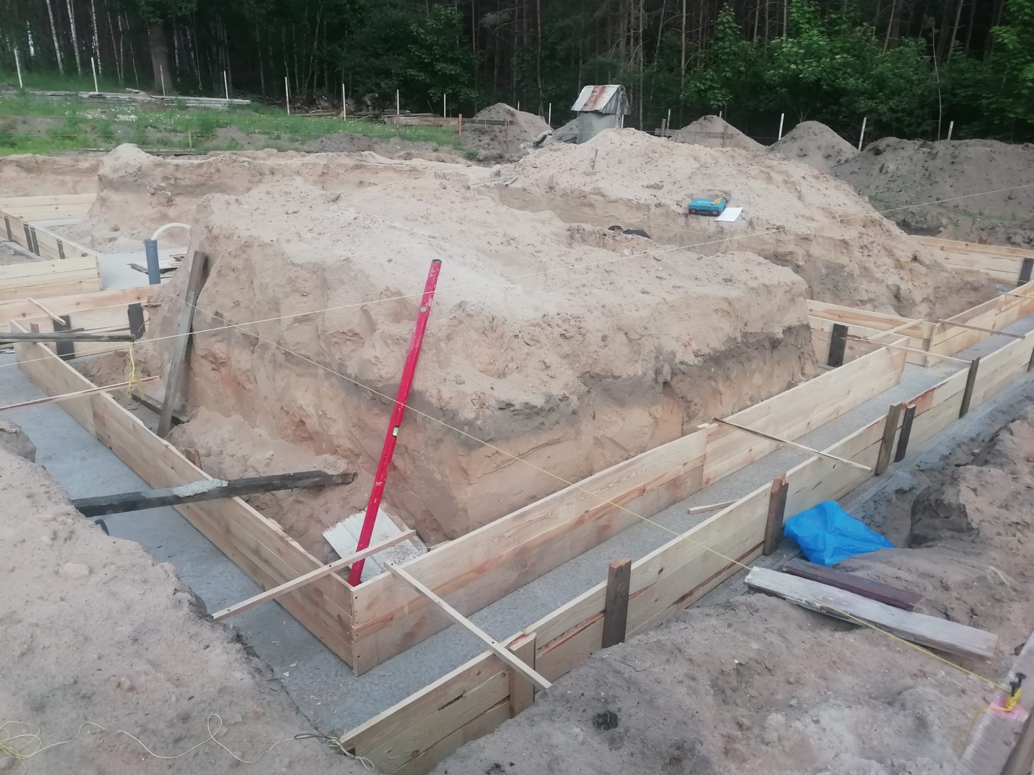 Fundamenty domu w trakcie budowy: drewniany szalunek, wykop, poziomica. Widoczne przygotowania do wylania betonu, teren budowy z ziemią i narzędziami.