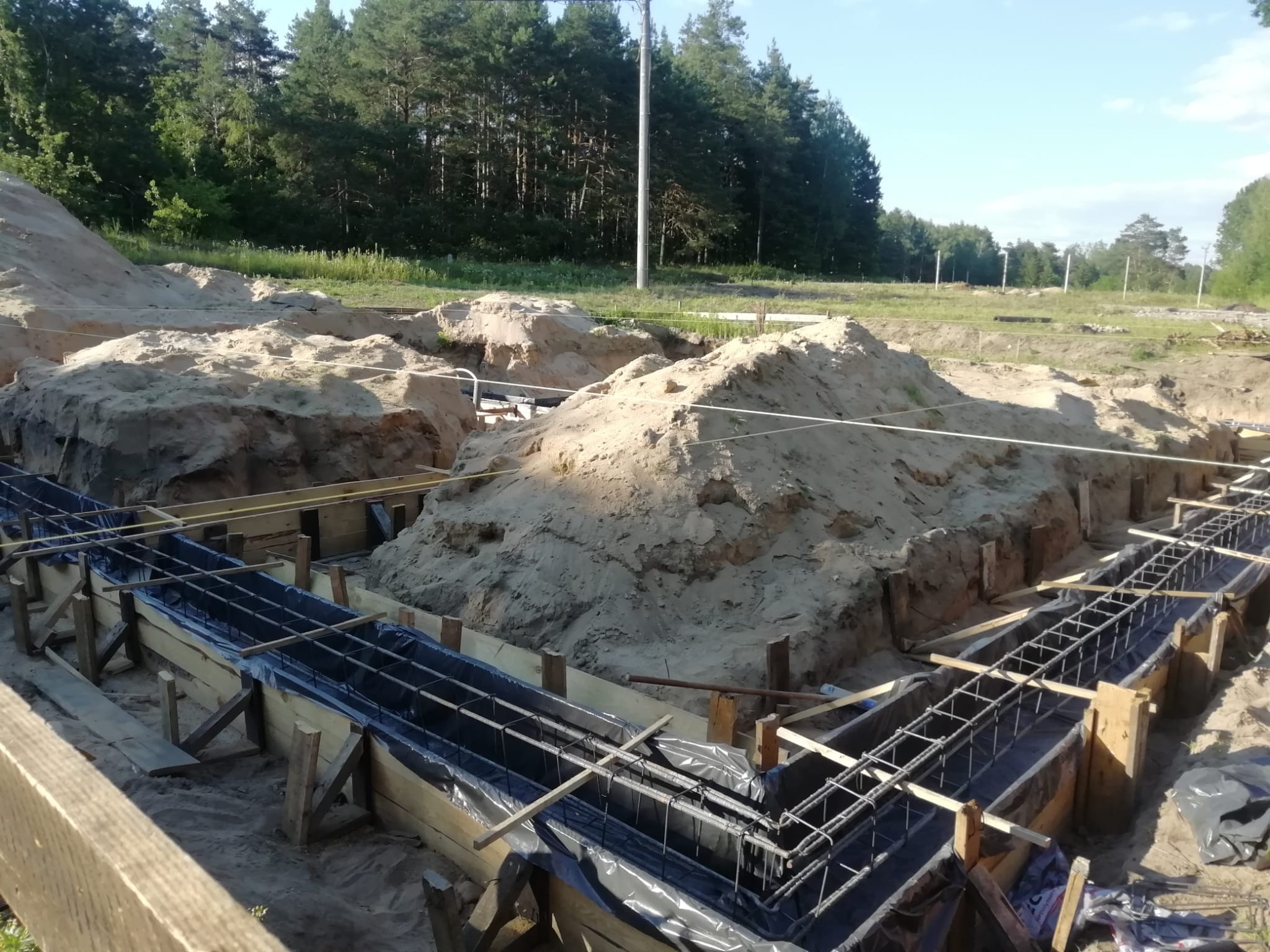Przygotowanie zbrojenia i szalunku pod fundament domu jednorodzinnego. Widoczne drewniane deski, stalowe pręty i folia ochronna. Wykop pod fundament.