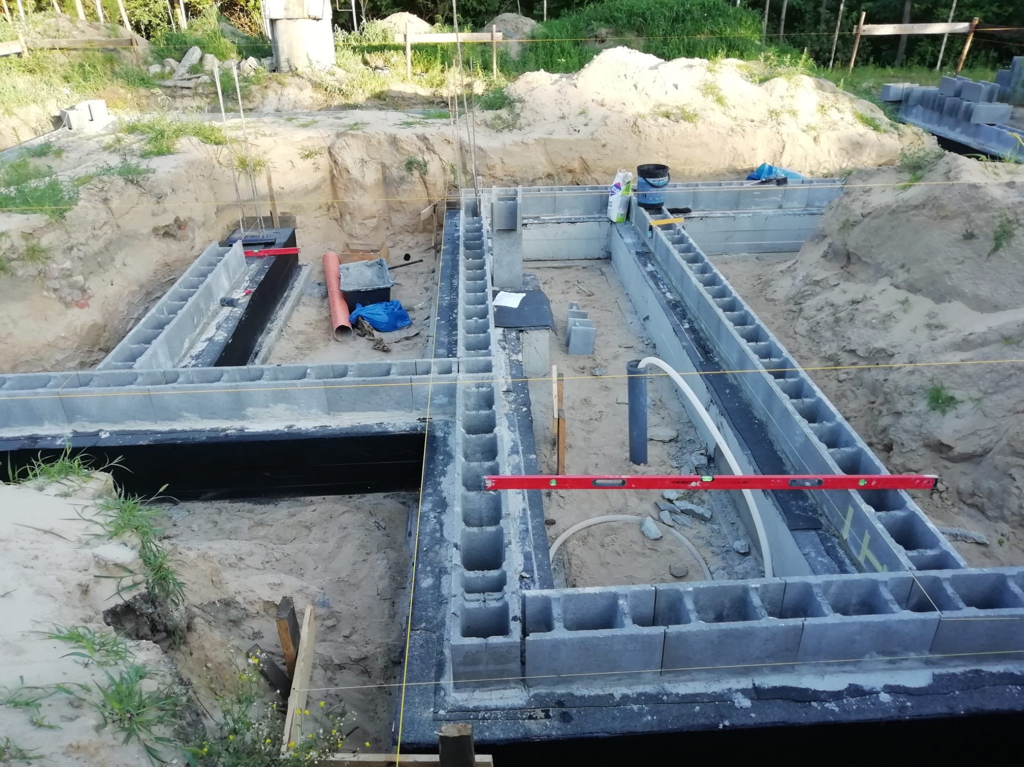 Fundamenty z bloczków betonowych w wykopie, poziomica, rury PCV, piasek, teren budowy. Widok z góry na wczesny etap budowy domu.