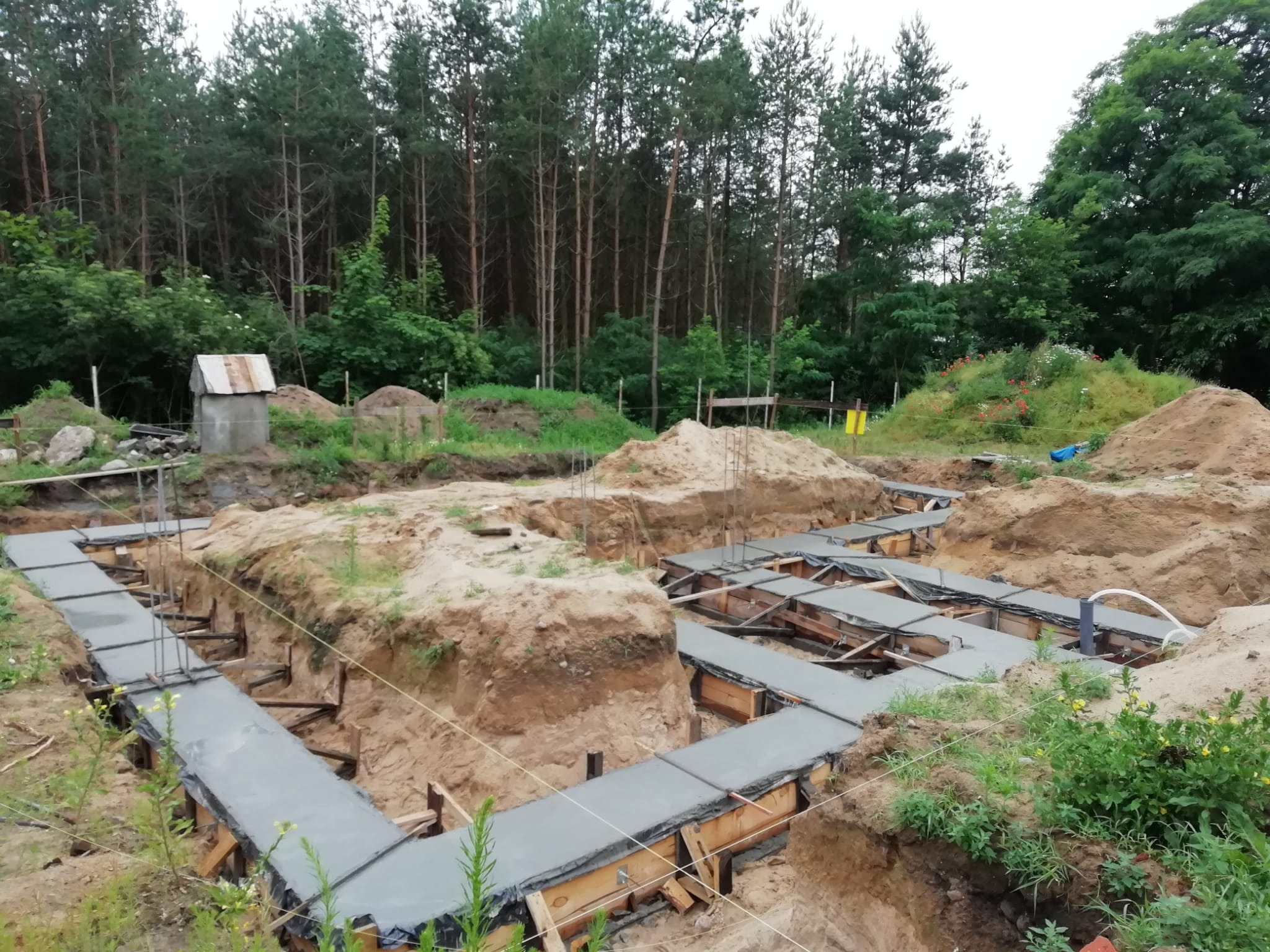 Zalane fundamenty pod budowę domu jednorodzinnego w Iłowo-Osada. Widoczne szalunki i wystające pręty zbrojeniowe. W tle las.