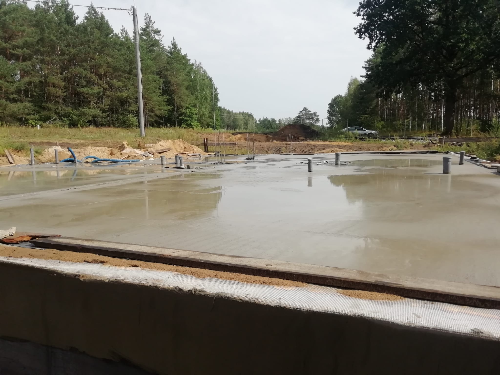 Zalana betonem płyta fundamentowa z wystającymi rurami instalacyjnymi na tle lasu i nieba. Widoczne szalunki i elementy konstrukcyjne.
