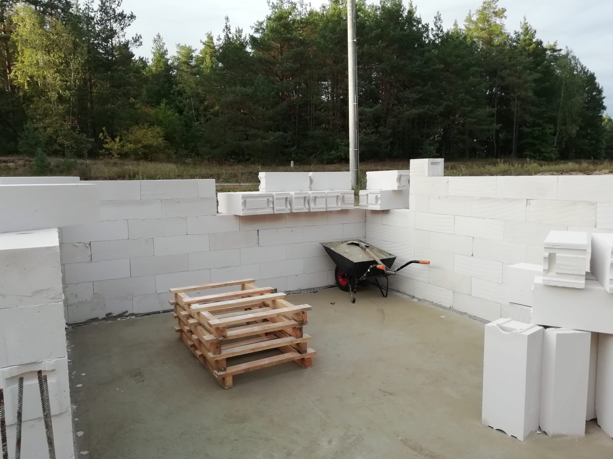 Budowa ścian z bloczków betonu komórkowego Ytong, widoczna paleta i taczka. Etap wznoszenia ścian fundamentowych na tle lasu.