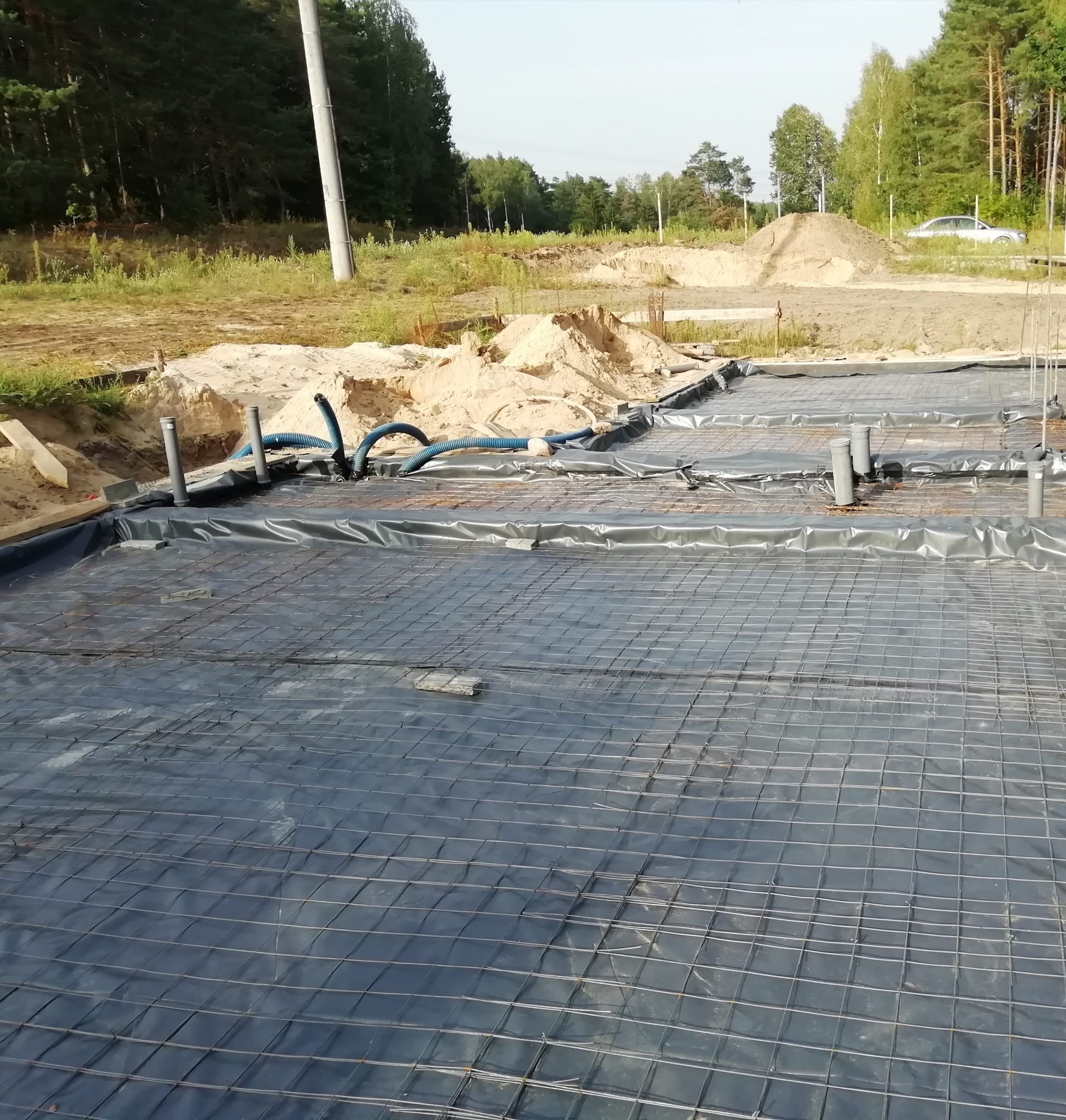 Zbrojenie fundamentów pod budowę domu, siatka stalowa na czarnej folii, wystające rury instalacyjne. W tle piasek i drzewa.