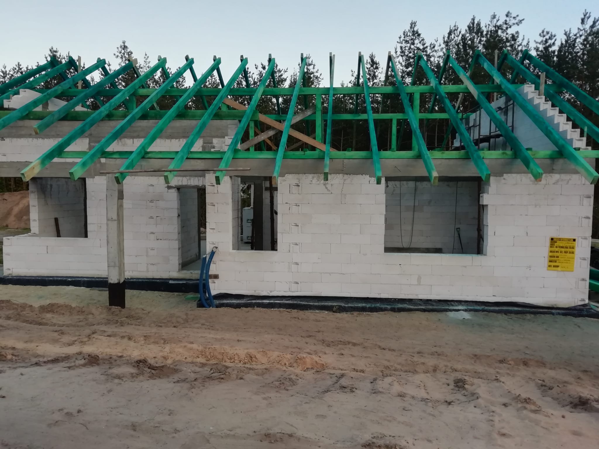 Budowa domu jednorodzinnego: ściany z białych bloczków, zielona konstrukcja dachu w trakcie montażu, widoczne okna i piasek wokół budynku. Tablica informacyjna na ścianie.