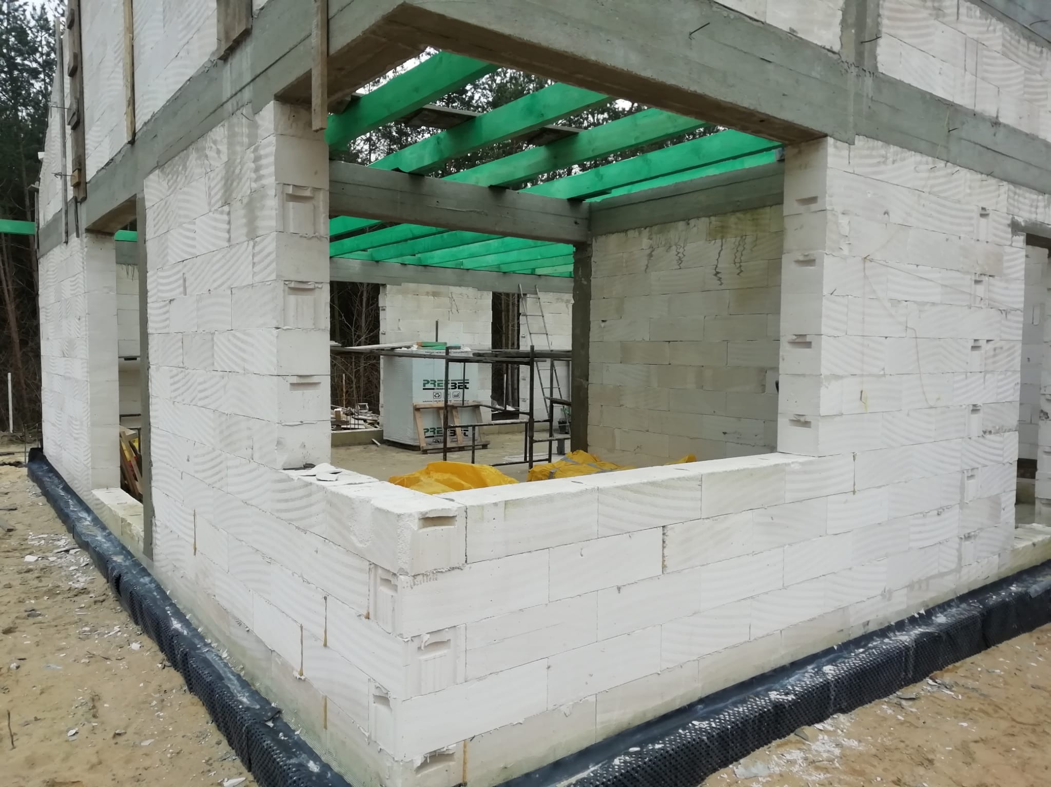 Budowa domu w stanie surowym otwartym: ściany z białych bloczków, drewniany strop pomalowany na zielono, widoczne rusztowania i materiały budowlane wewnątrz.