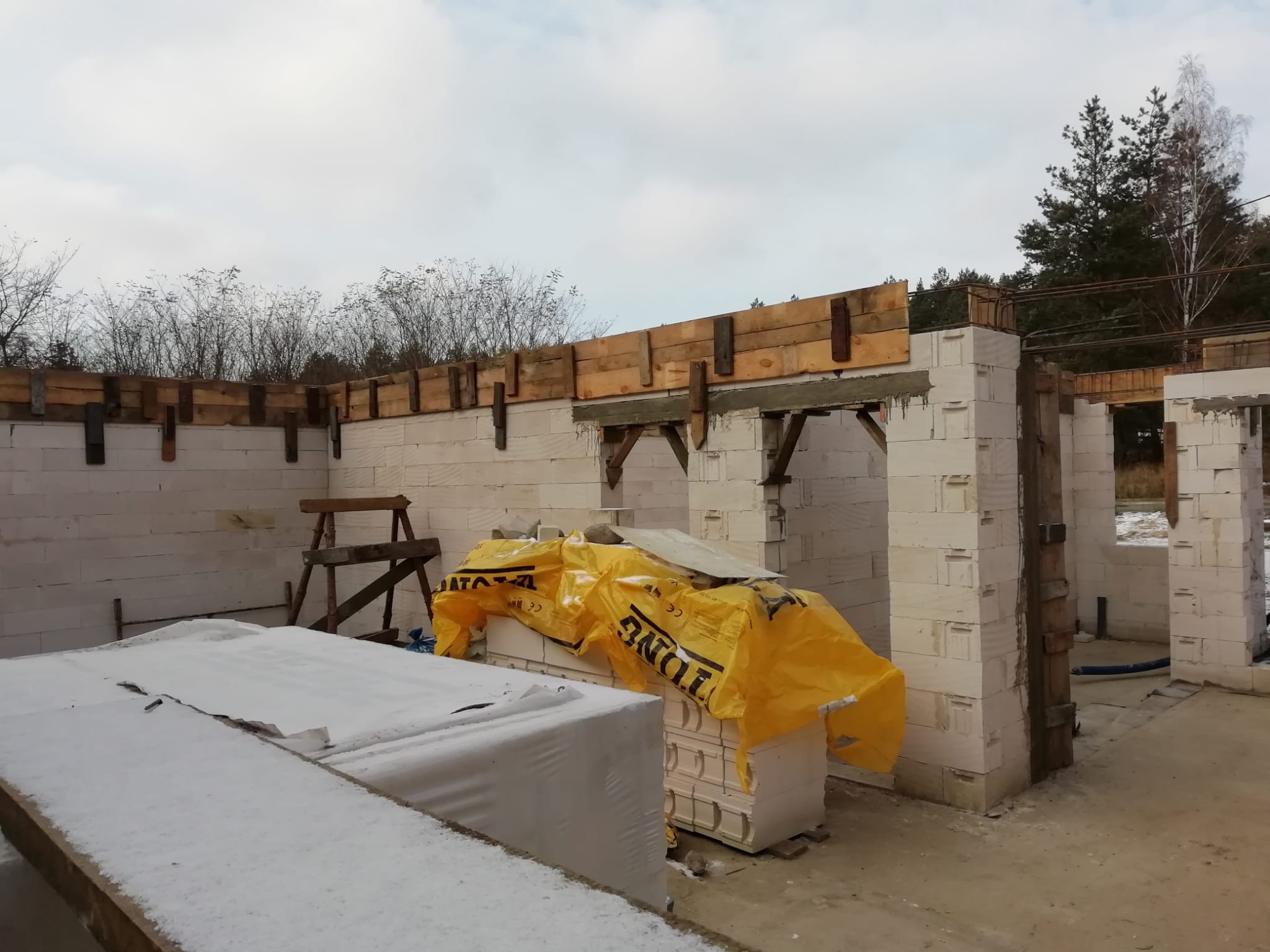 Wznoszenie ścian z bloczków betonu komórkowego Ytong. Widoczne drewniane szalunki nad otworami okiennymi i drzwiowymi. Część bloczków przykryta żółtą folią.