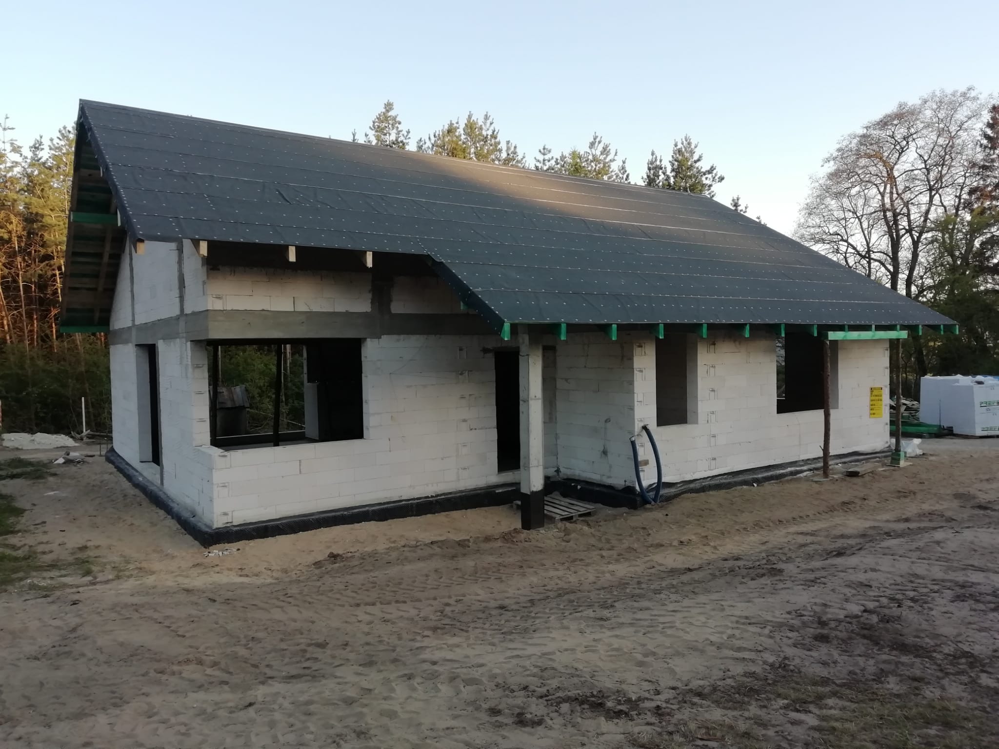 Surowy stan domu jednorodzinnego z szarym dachem, widoczne pustaki i przygotowane otwory okienne, otoczenie piaszczyste, drzewa w tle.