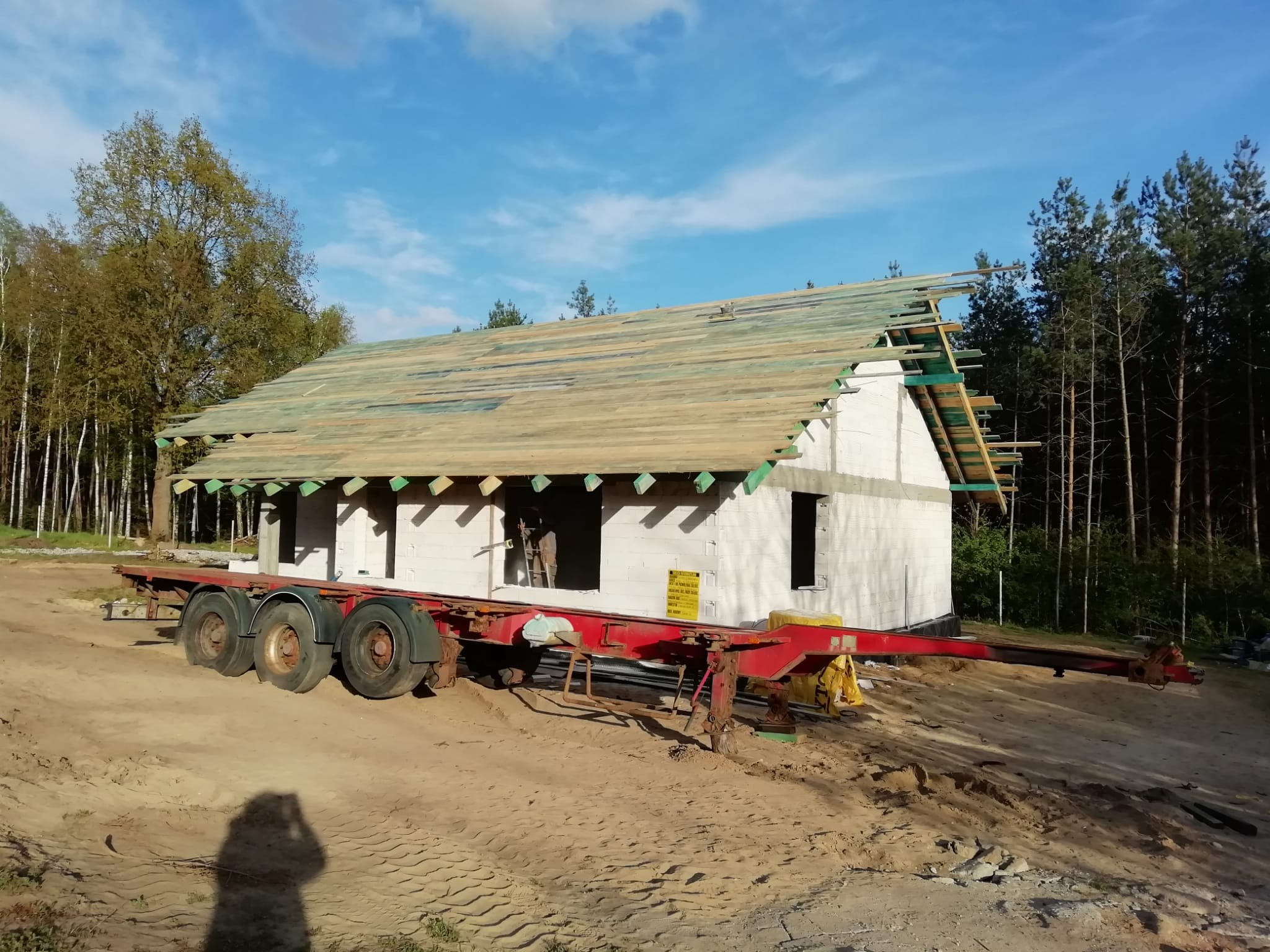 Niedokończony dom z białych bloczków na czerwonej platformie transportowej, z częściowo ułożonym drewnianym dachem na tle lasu i błękitnego nieba.