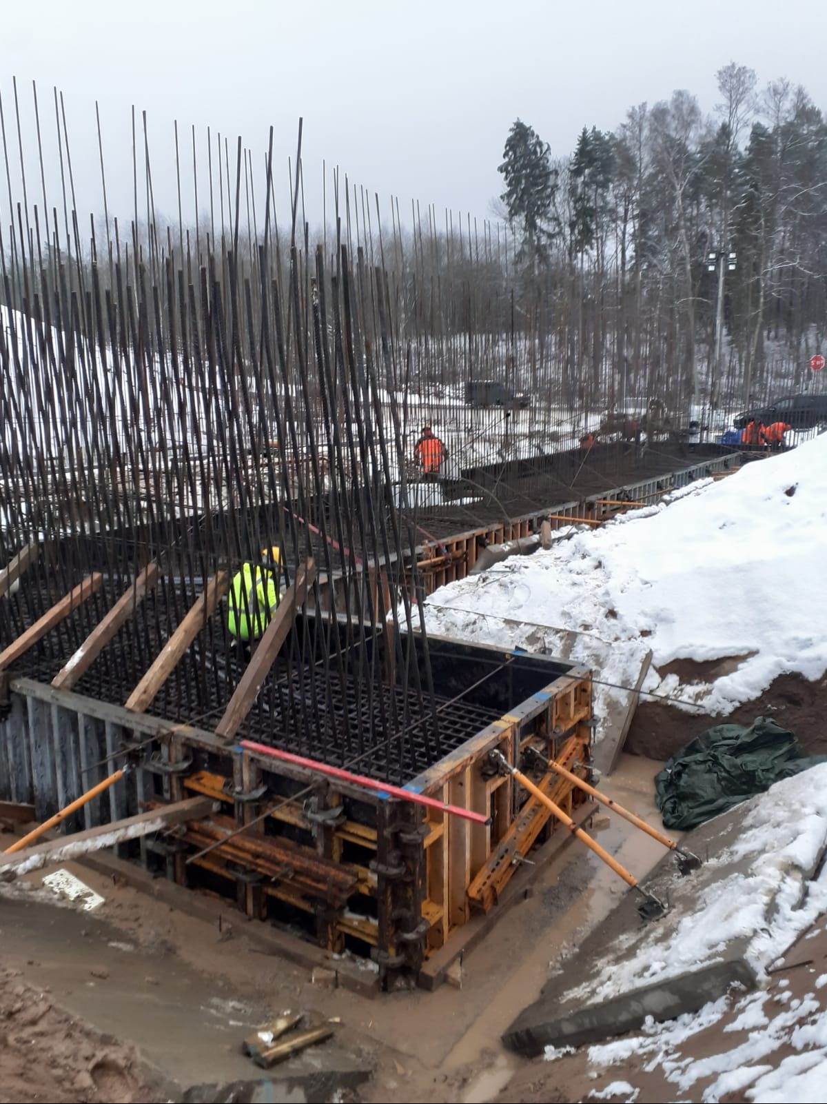Zbrojenie i szalunek pod fundamenty mostu w zimowej scenerii. Widoczne pręty zbrojeniowe, drewniane podpory i pracownicy w odblaskowych ubraniach.