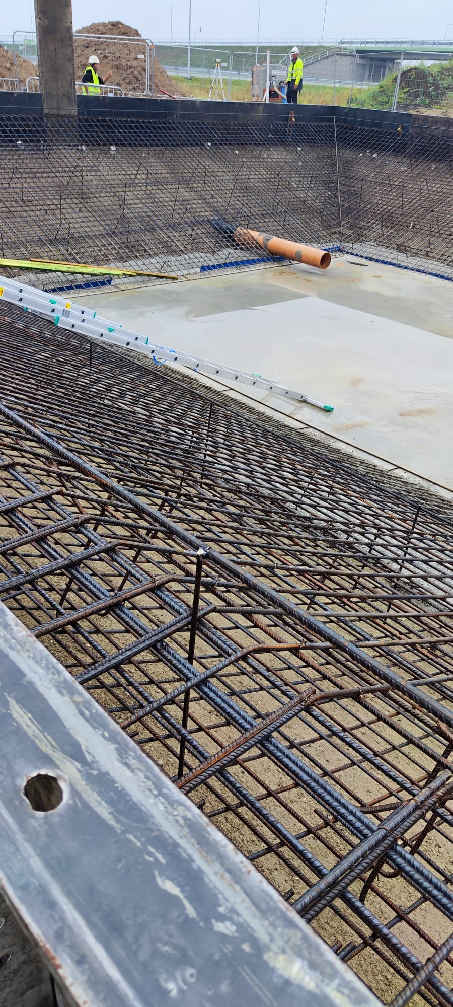 Zbrojenie fundamentów pod budowę, widoczne pręty zbrojeniowe ułożone w siatkę, częściowo zalane betonem. Na górze widoczni pracownicy w kaskach.