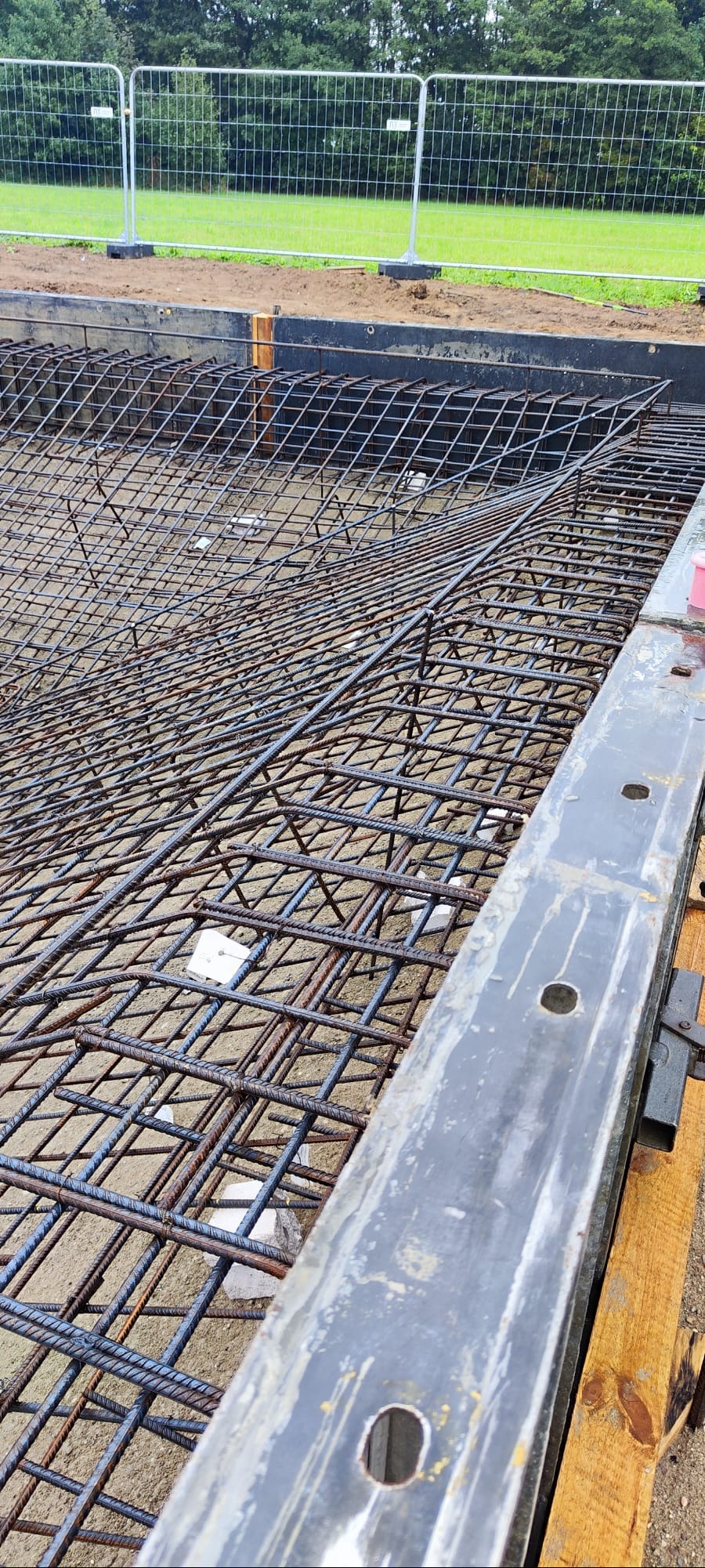 Zbrojenie fundamentów z siatki stalowej, widoczne białe podkładki dystansowe. Szalunek z metalowych belek. W tle trawa i ogrodzenie.