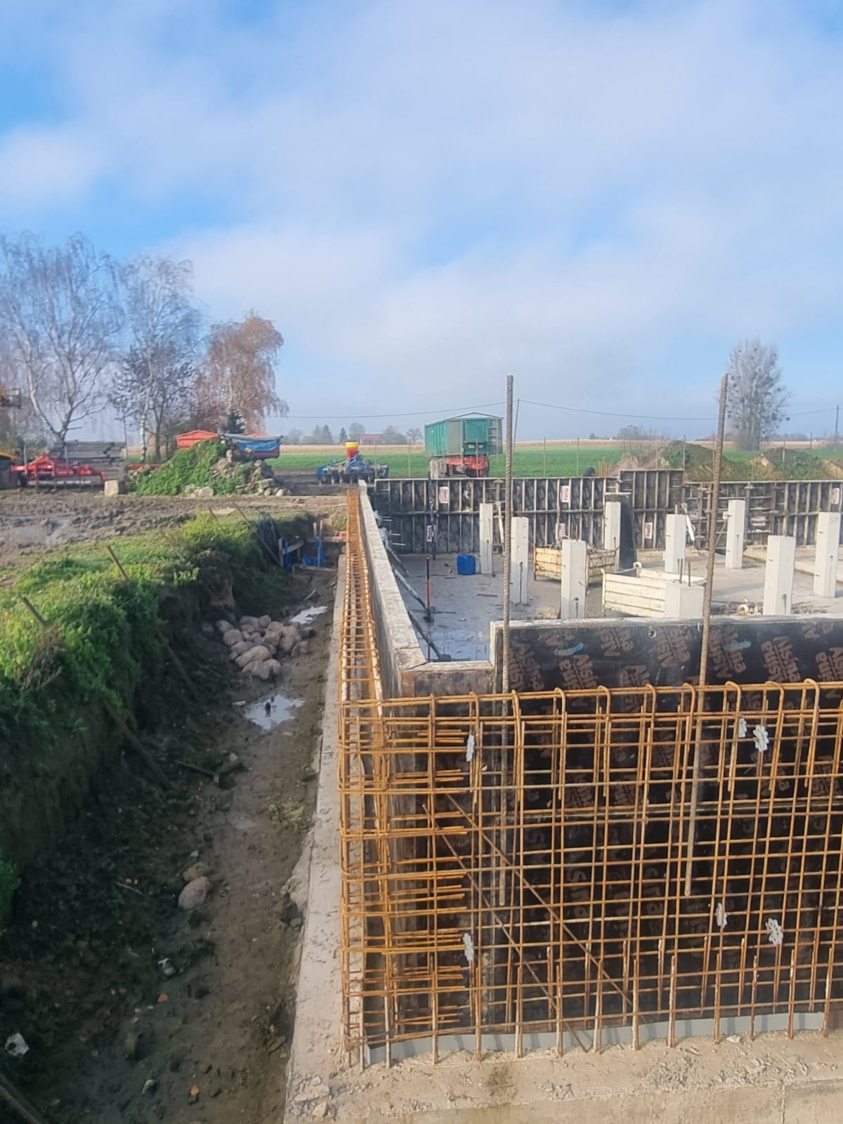Zbrojenie fundamentów pod budynek jednorodzinny, widoczne szalunki i stalowe pręty. W tle krajobraz wiejski z maszynami rolniczymi i błękitne niebo.