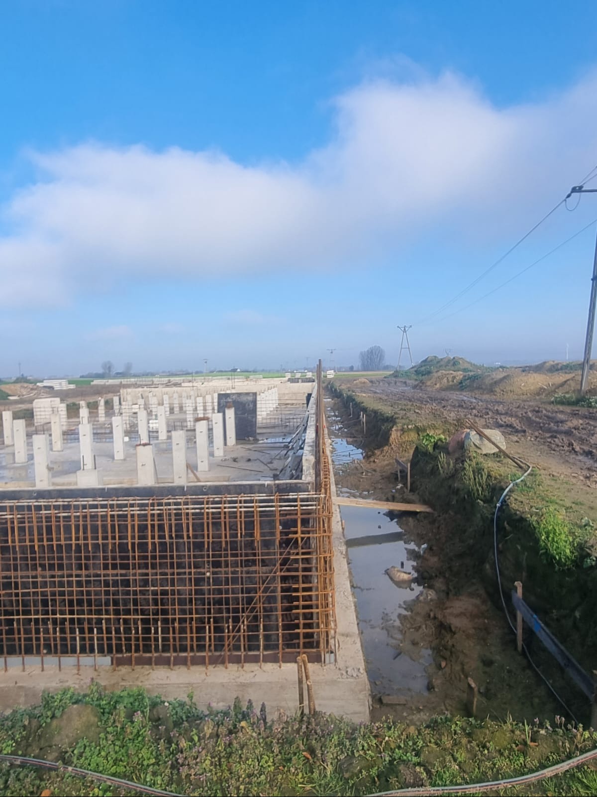 Fundamenty budynku w trakcie budowy: zbrojenie, betonowe słupy i ściany, ziemia. Widok na plac budowy w słoneczny dzień.