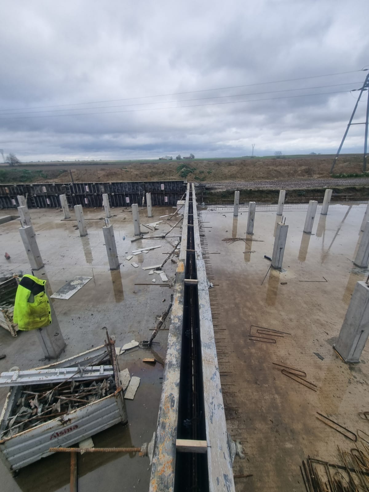 Wylany fundament z betonowymi słupami i szalunkiem w trakcie budowy, widok z góry na mokrą powierzchnię placu budowy w pochmurny dzień.
