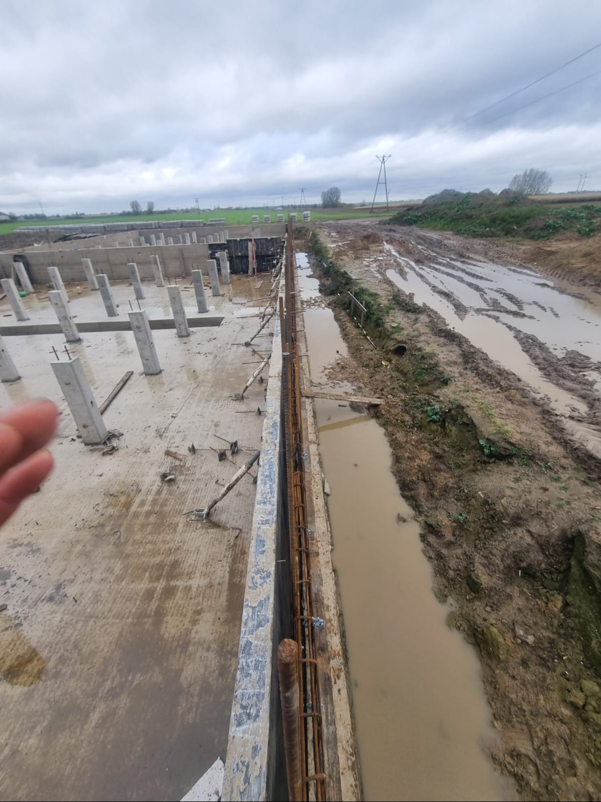 Fundamenty budynku w trakcie budowy: betonowe słupy, zbrojenie, szalunki. Teren budowy błotnisty, pochmurne niebo w tle. Widok z góry, częściowo widoczna dłoń.