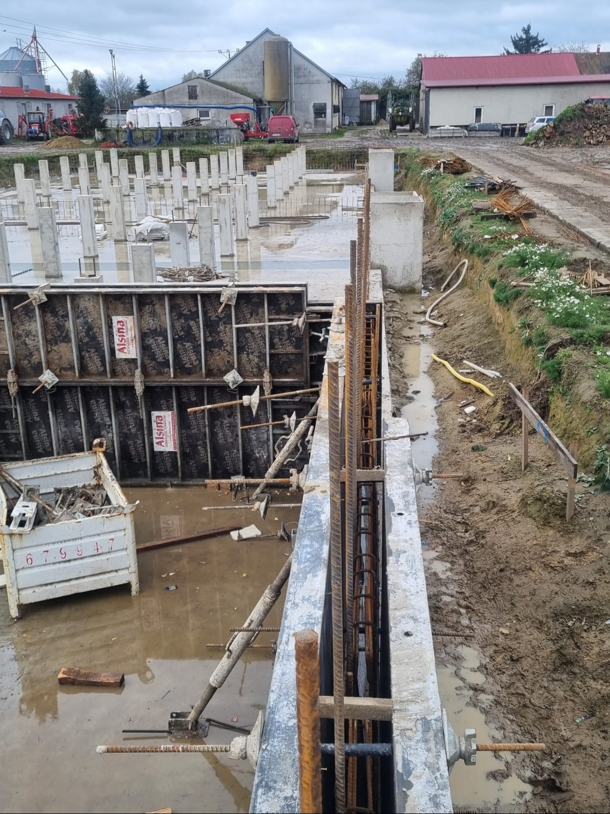 Fundamenty budynku w trakcie budowy: szalunki, zbrojenie, zalane wodą. Widoczne stemple i elementy konstrukcyjne. W tle budynki gospodarcze i sprzęt rolniczy.