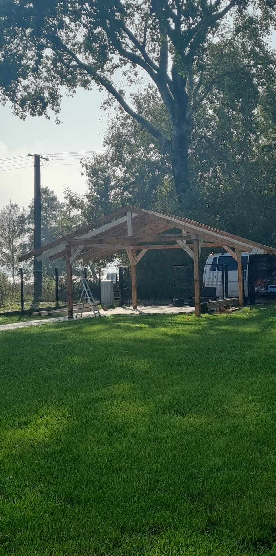 Drewniana konstrukcja dachu w trakcie budowy, widoczna drabina, trawnik i drzewa w tle. Ujęcie w słoneczny dzień.