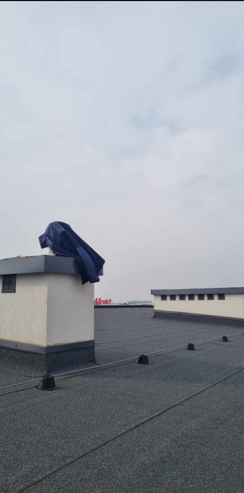 Płaski dach pokryty papą z kominem i elementami odgromowymi. Na kominie leży niebieska plandeka. W tle budynek z napisem 'Markt'. Pochmurne niebo.