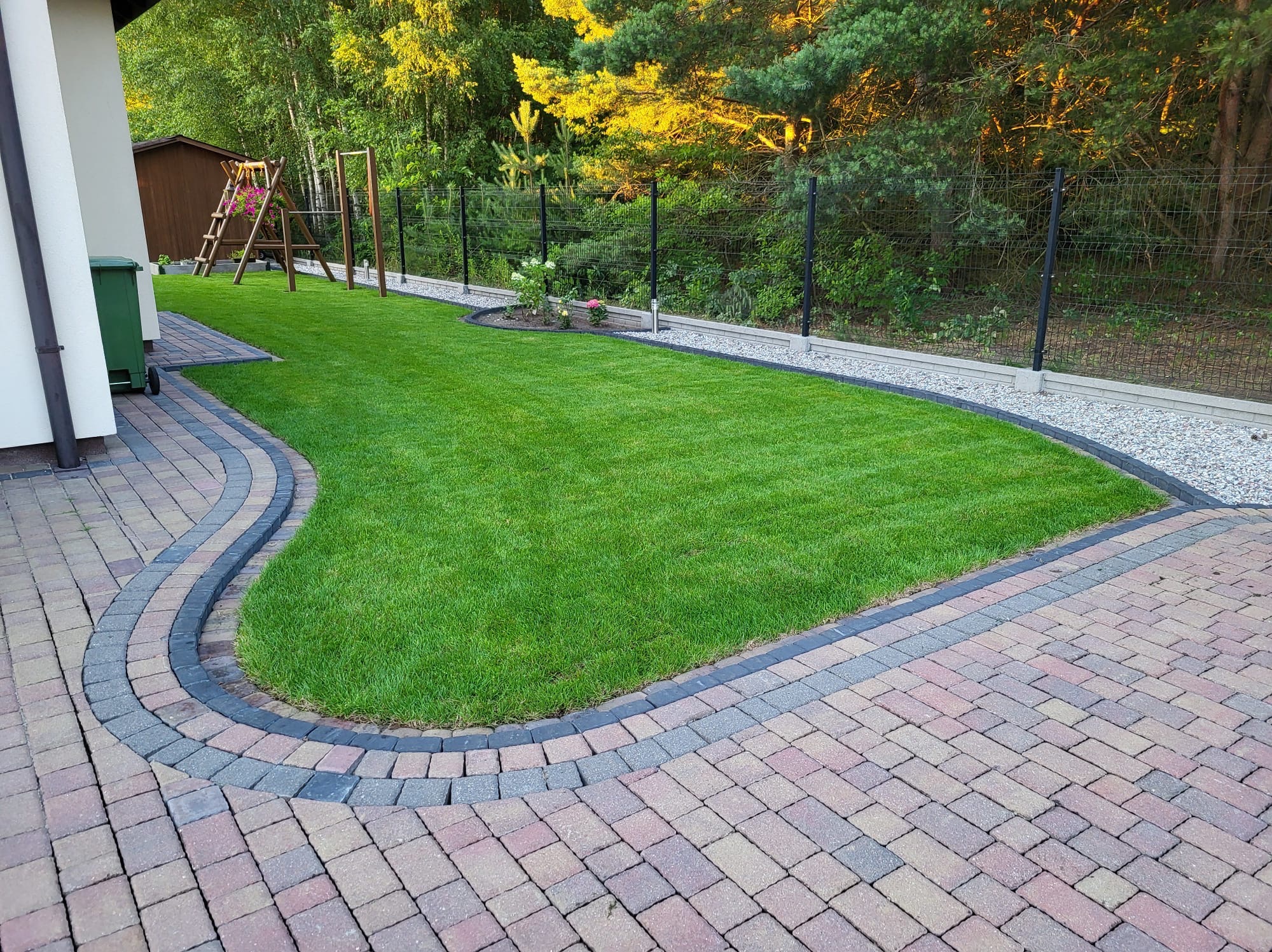 Zadbany trawnik z kostką brukową i obrzeżami, widok z góry. Na trawniku mały klomb z kwiatami. W tle siatka ogrodzeniowa i drzewa.
