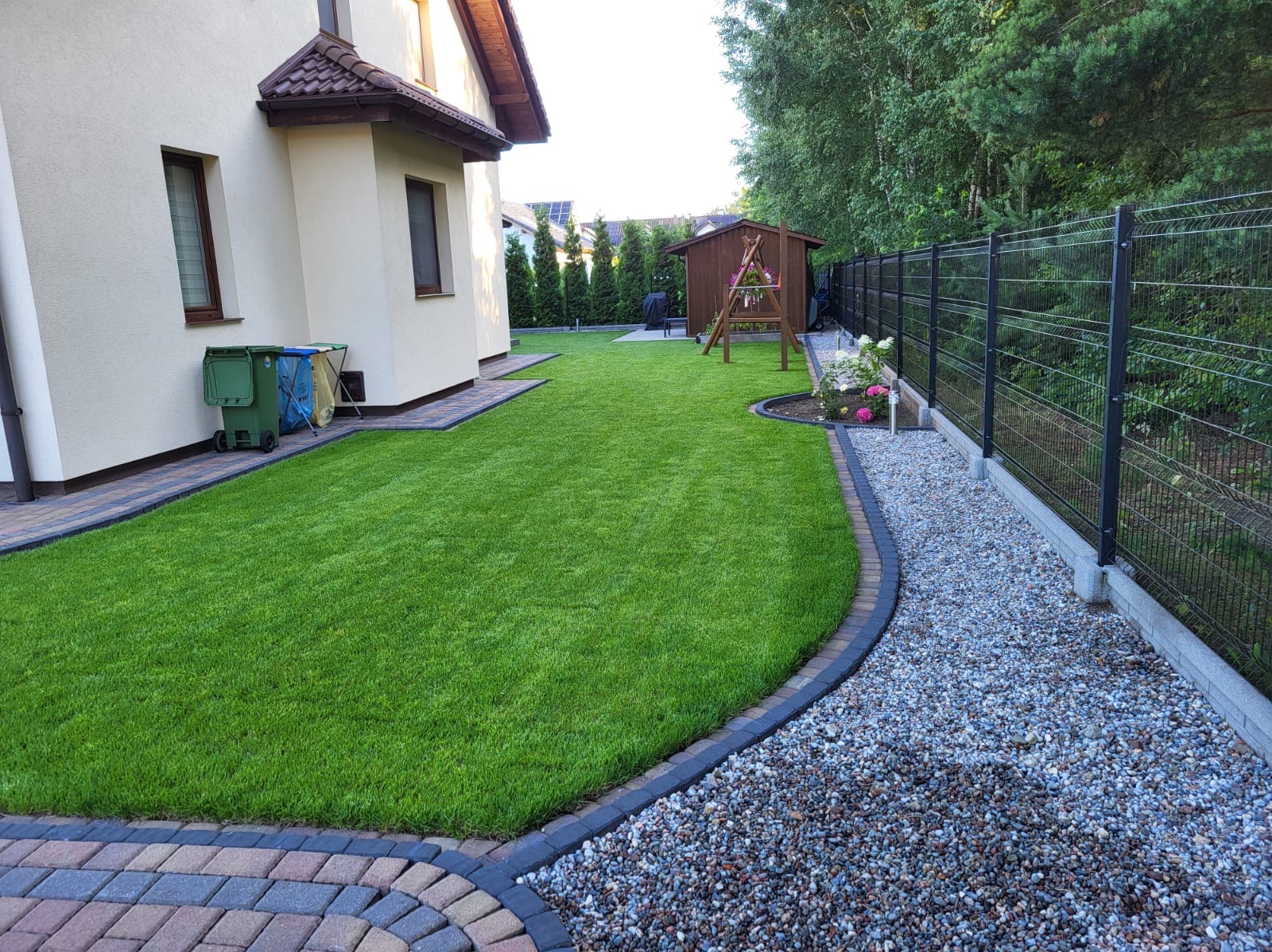 Zadbany trawnik z rolki, obrzeża z kostki, szara rabata z kamienia wzdłuż ogrodzenia panelowego. W tle domek narzędziowy i wysokie tuje.