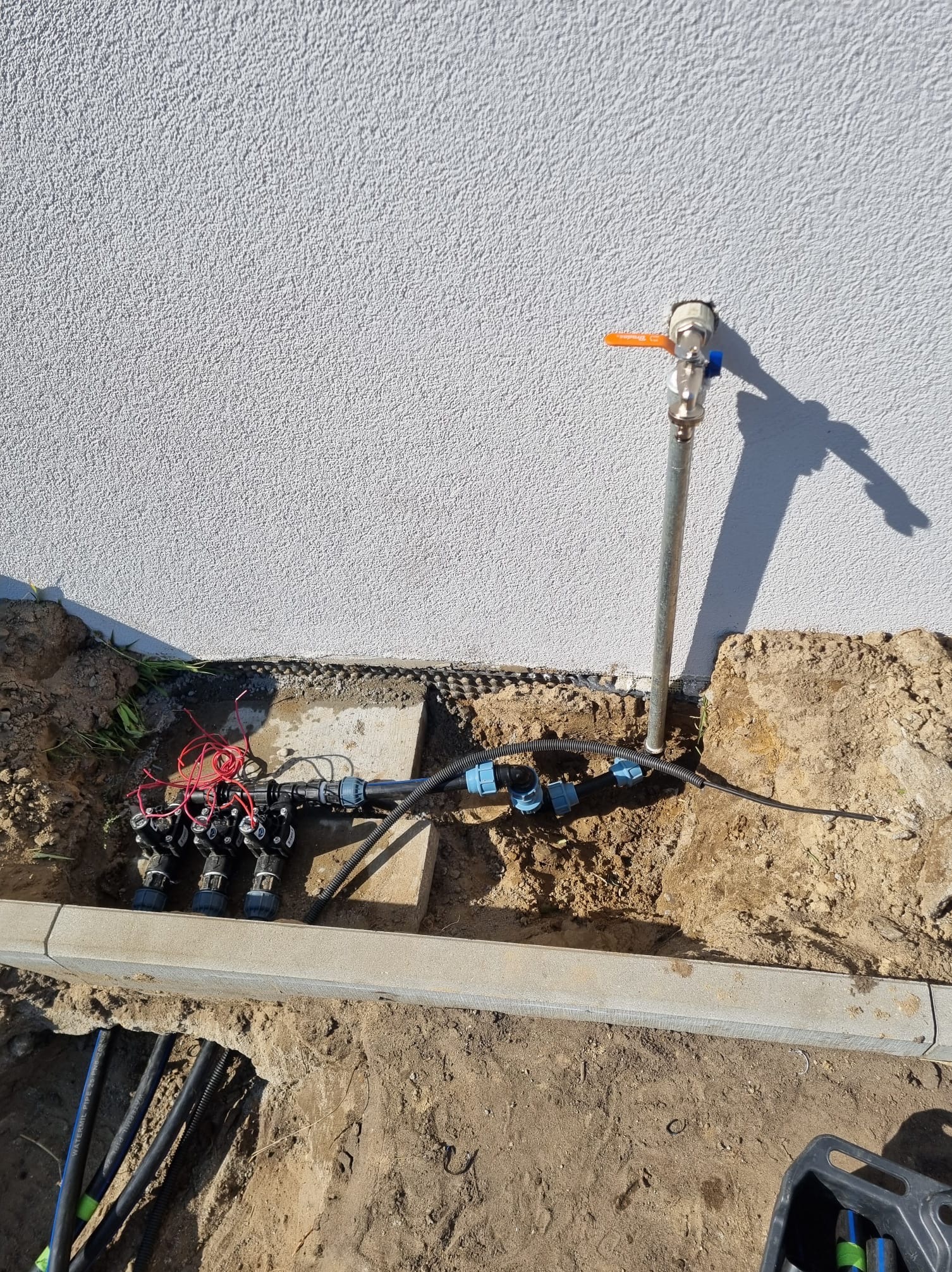 Instalacja systemu nawadniania w ogrodzie: widoczne rury, zawory i przyłącze wodne przy ścianie budynku. Wykopane rowy, ziemia i narzędzia ogrodnicze.