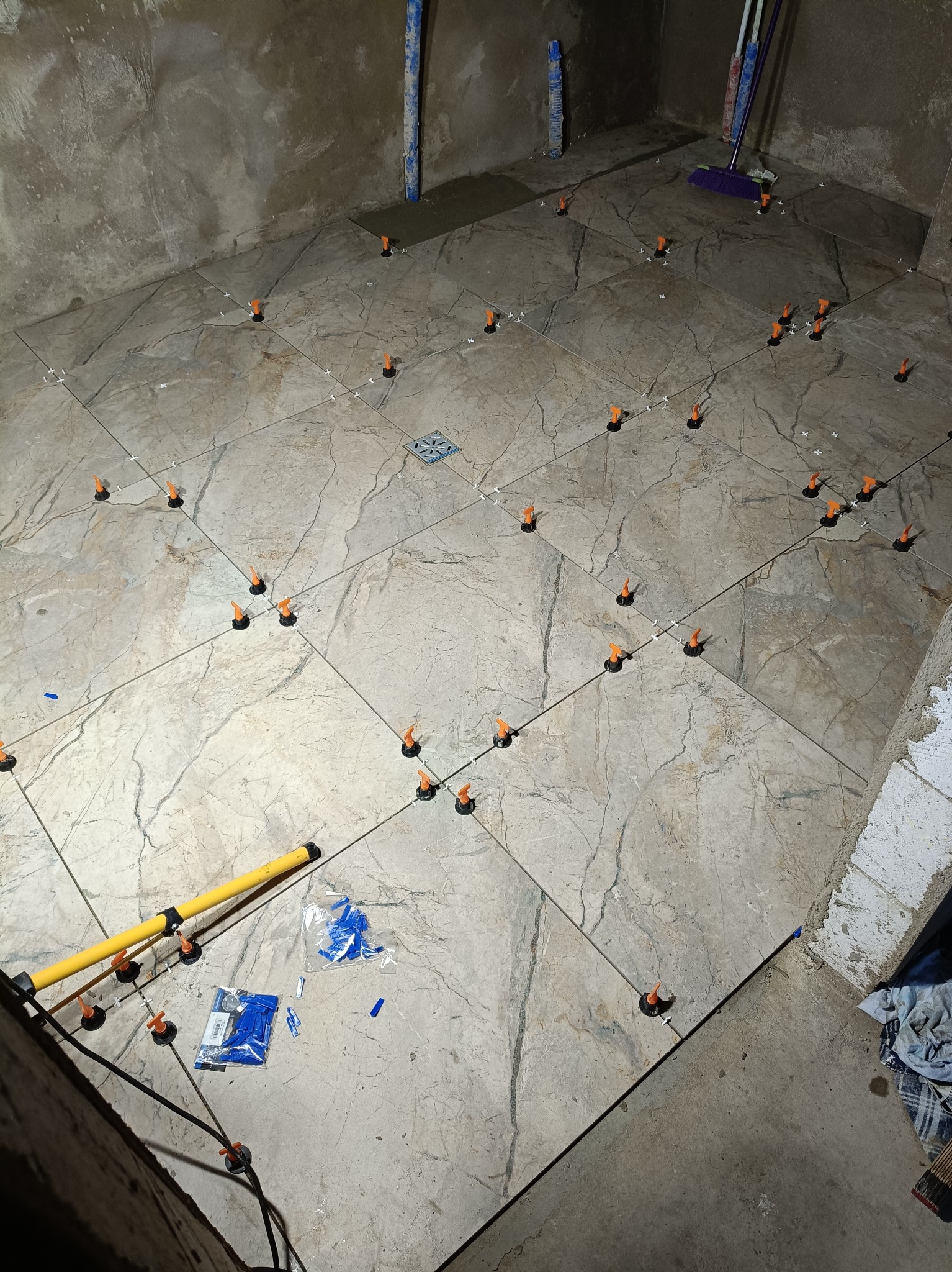 Układanie płytek podłogowych z użyciem systemu poziomowania. Widoczne płytki imitujące marmur, krzyżyki dystansowe i narzędzia glazurnicze. Etap prac wykończeniowych w łazience.