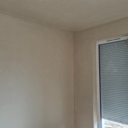Jakub Porzyczek - Pokój po nałożeniu gładzi gipsowej, widoczne ślady po pracach na podłodze i wystający kabel z sufitu. Okno z roletami w tle.