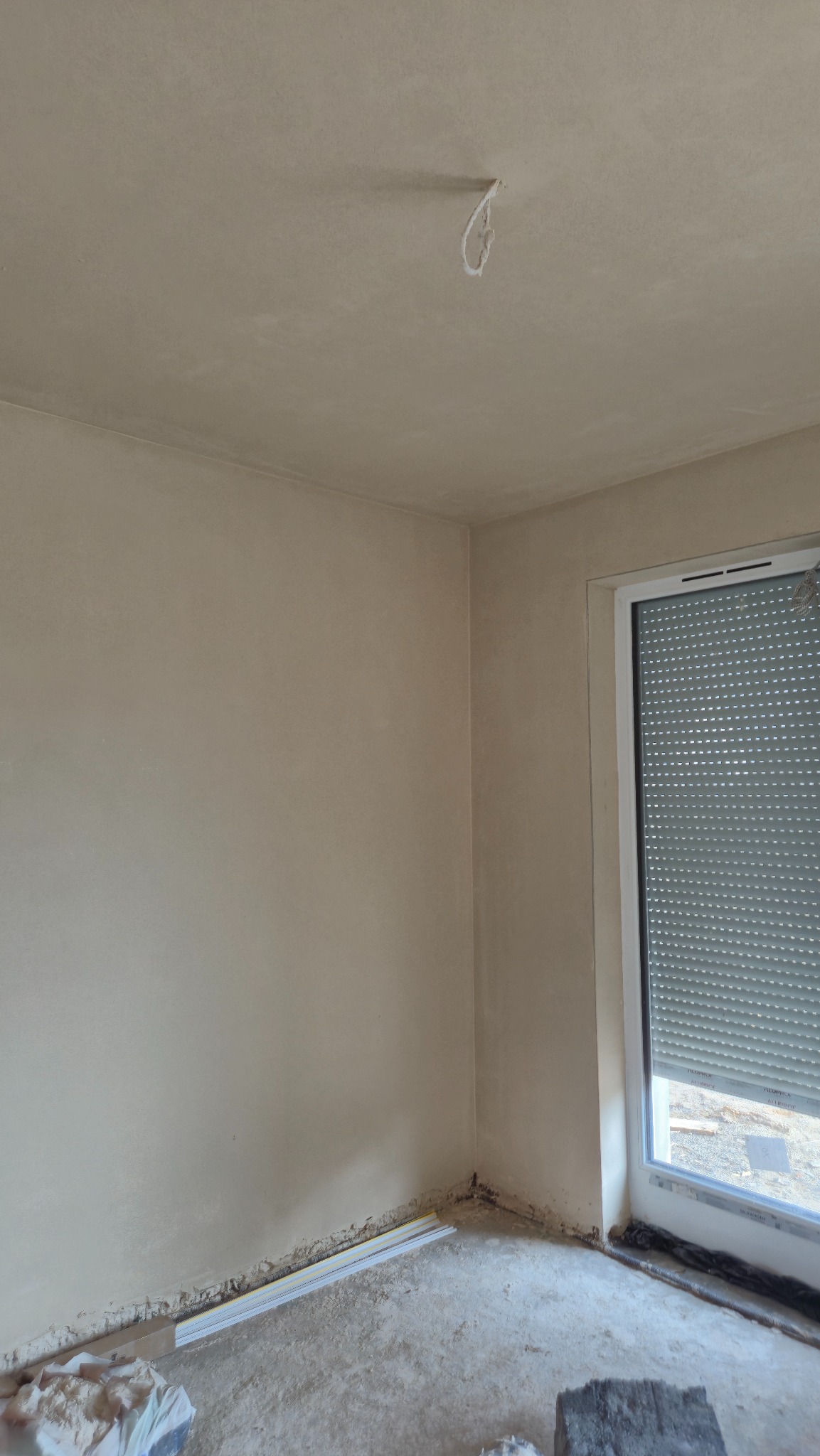 Pokój po nałożeniu gładzi gipsowej, widoczne ślady po pracach na podłodze i wystający kabel z sufitu. Okno z roletami w tle.