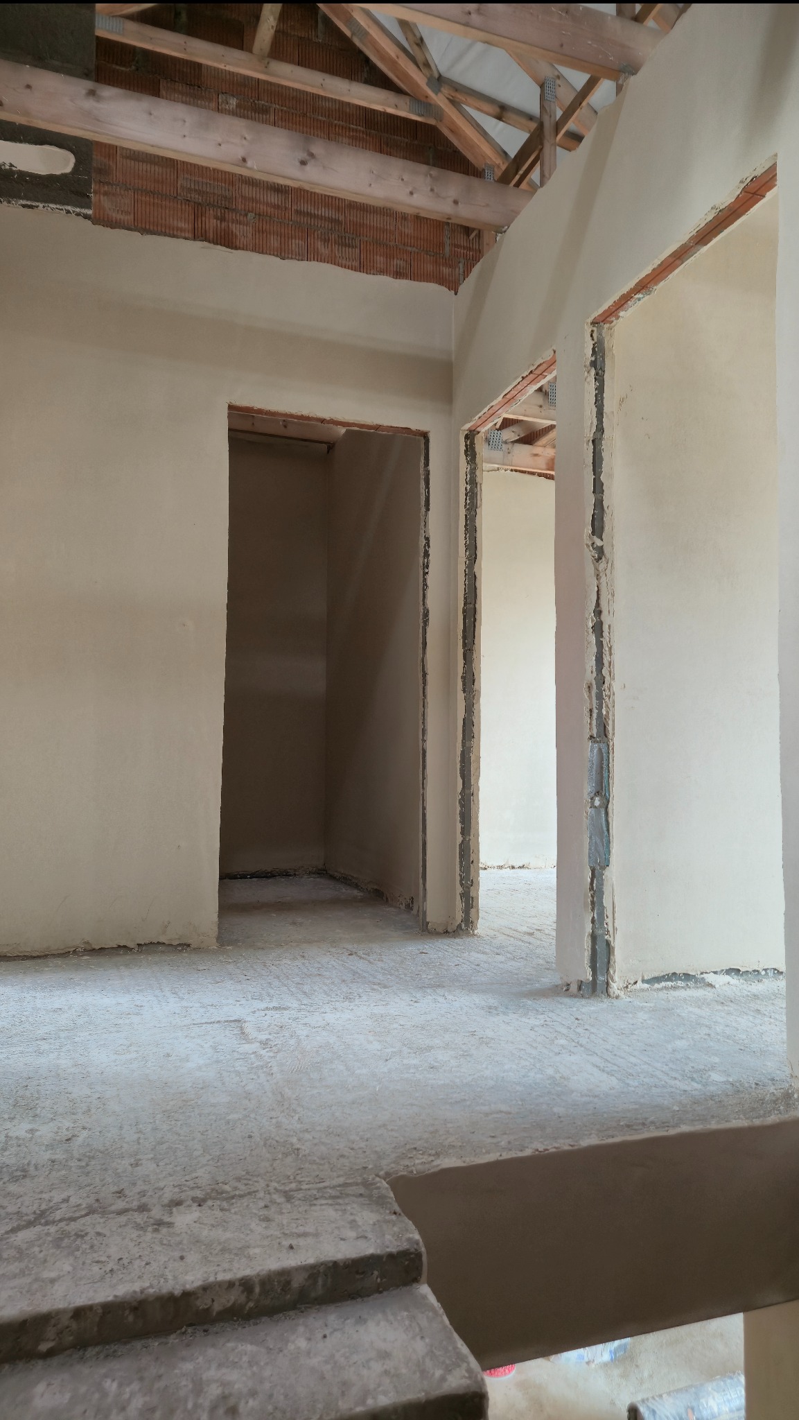 Wnętrze w stanie surowym po tynkowaniu ścian. Widoczne otwory drzwiowe, surowy strop i betonowa podłoga. Ściany pokryte jasnym tynkiem.