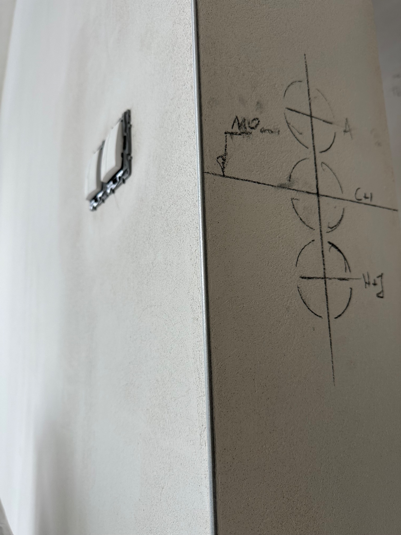 Ściana z zaznaczonymi punktami instalacji elektrycznej i gniazdkiem. Widoczne odręczne oznaczenia i linie pomocnicze. Szary odcień ściany.