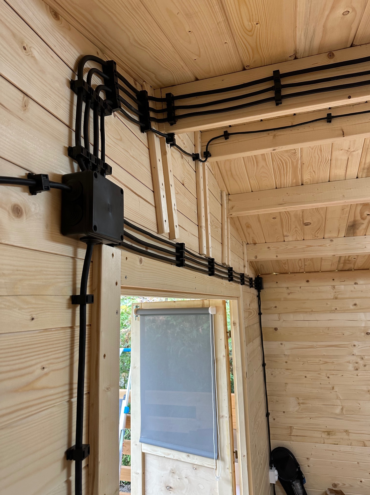 Czarna instalacja elektryczna natynkowa w drewnianym domku, kable prowadzone w listwach, puszka rozgałęźna, widok na drzwi z roletą.