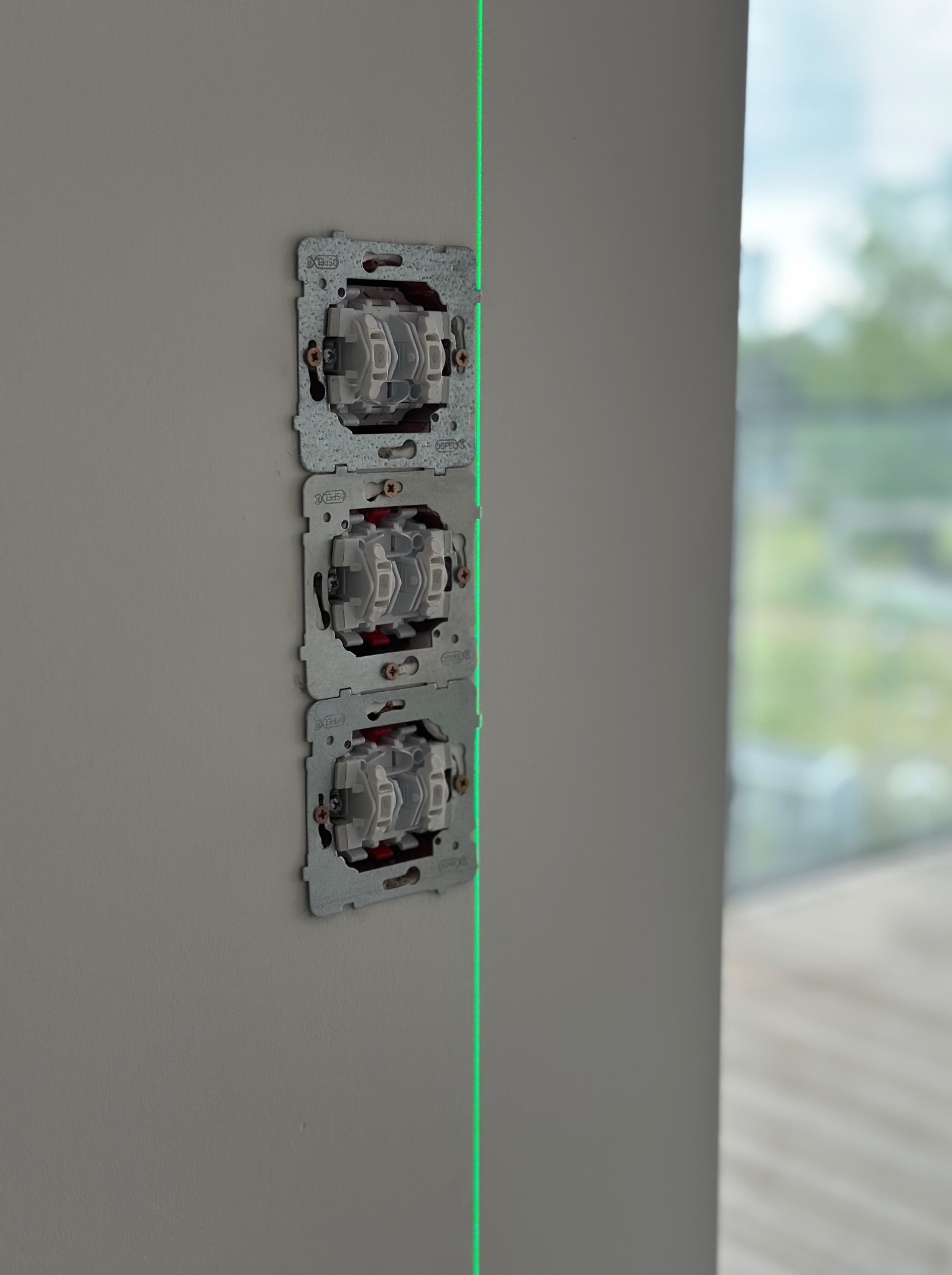 Potrójny zestaw podtynkowych puszek elektrycznych Ospel, zamontowanych pionowo na szarej ścianie, wyrównanych laserowo. Widok z boku, z oknem w tle.