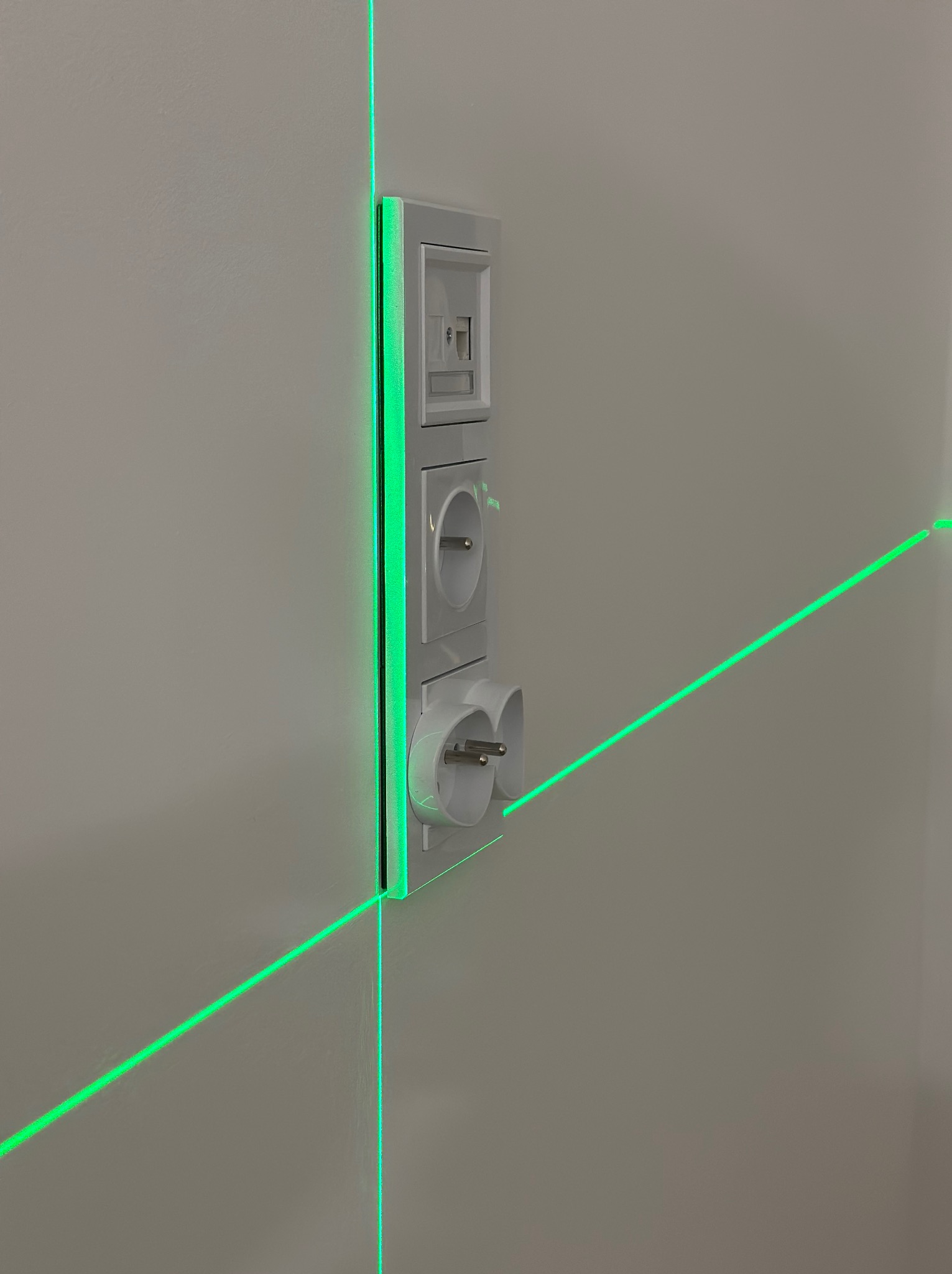 Białe gniazdka elektryczne i gniazdo antenowe na ścianie, wyznaczanie poziomu laserem krzyżowym z zieloną wiązką. Precyzyjny montaż osprzętu elektrycznego.