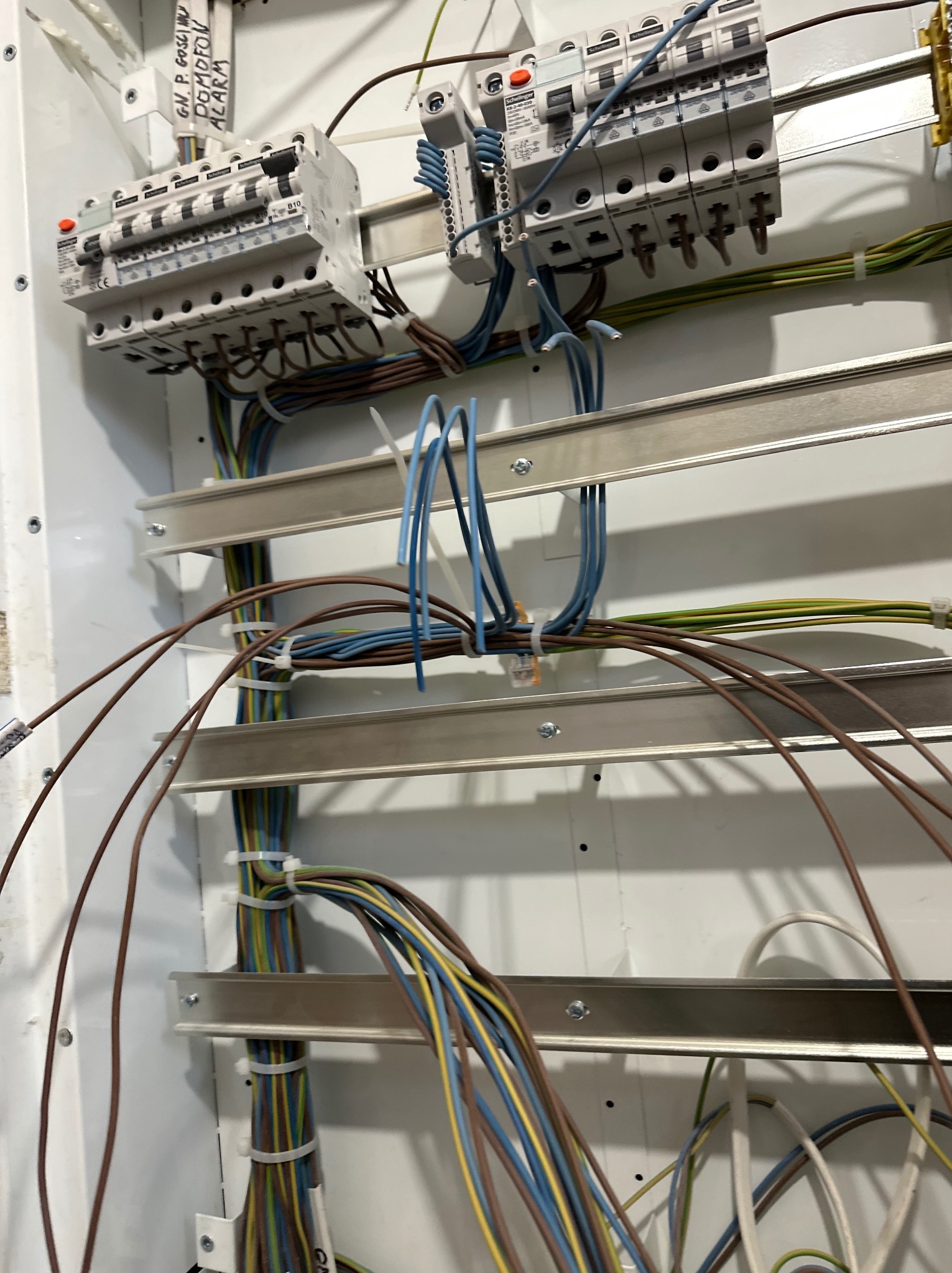 Rozdzielnica elektryczna z bezpiecznikami i uporządkowanymi przewodami w kolorach: brązowym, niebieskim, żółto-zielonym. Widoczne szyny montażowe i oznaczenia na obudowie.