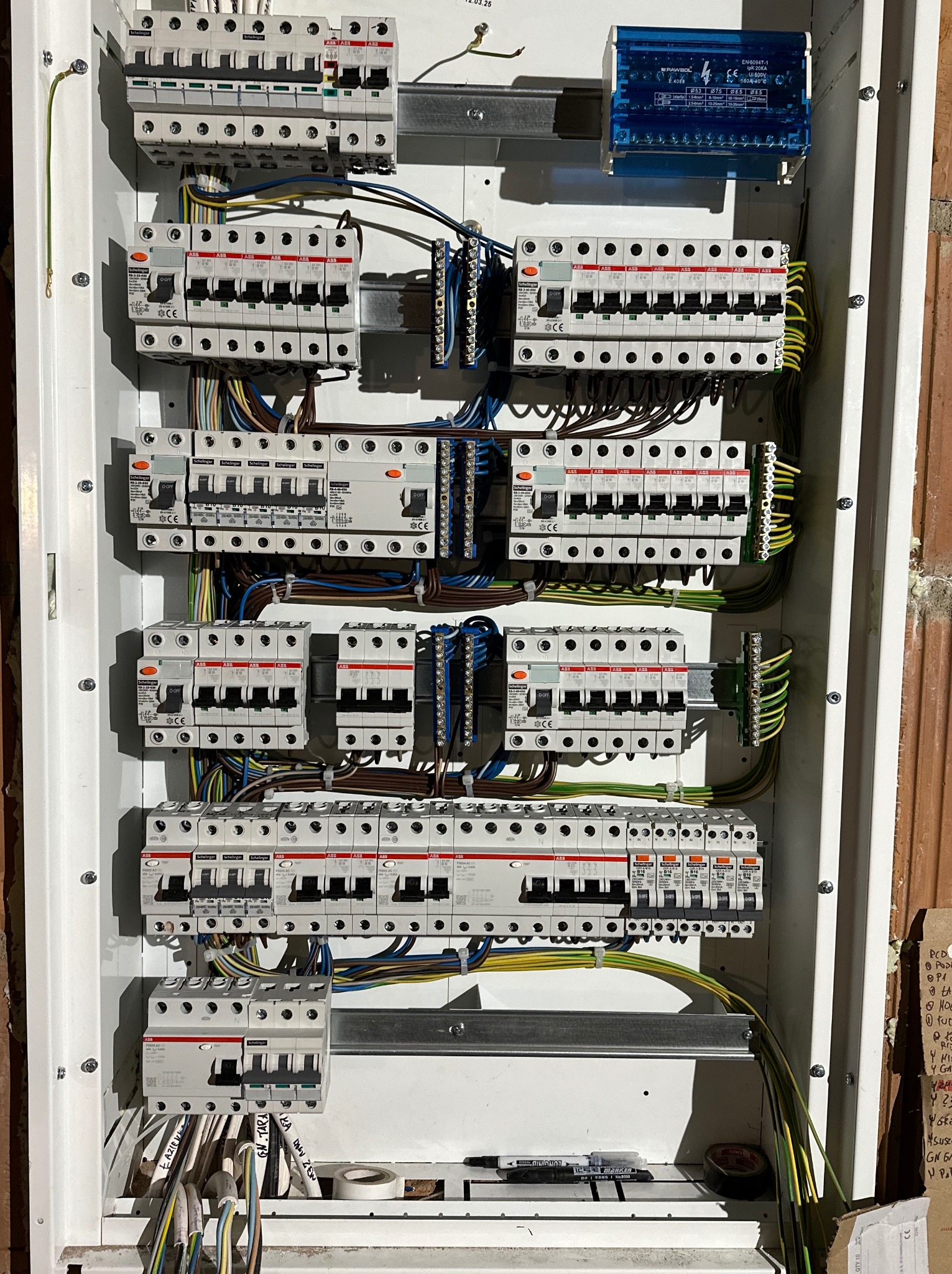 Rozdzielnica elektryczna z zamontowanymi bezpiecznikami i okablowaniem, widok z bliska. Elementy instalacji elektrycznej w białej obudowie na tle ceglanej ściany.