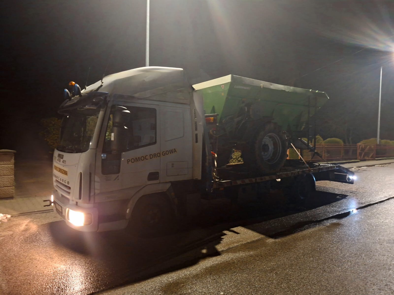 Biała laweta pomocy drogowej przewozi zielony rozsiewacz na niskopodwoziowej platformie w nocnych warunkach oświetleniowych.