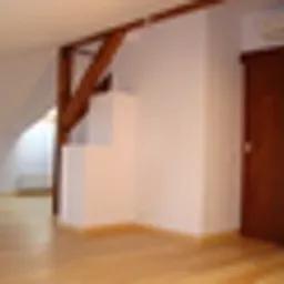 Puste, jasne wnętrze poddasza z drewnianą podłogą i widocznymi elementami konstrukcyjnymi dachu. Ściany pomalowane na biało, widoczne schody obłożone białymi płytami. Zamknięte, brązowe drzwi po...