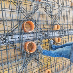 DREBEKO - Zbrojenie fundamentów z rurami kanalizacyjnymi na drewnianym szalunku, widok z góry. Fragment nóg w jeansach i butach roboczych na konstrukcji.