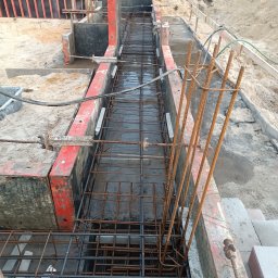 DREBEKO - Zbrojenie fundamentów w szalunkach, widok z góry. Stalowe pręty tworzą siatkę, częściowo zalane wodą. Czerwone szalunki z widocznymi śladami użytkowania. Piasek i ziemia w tle.